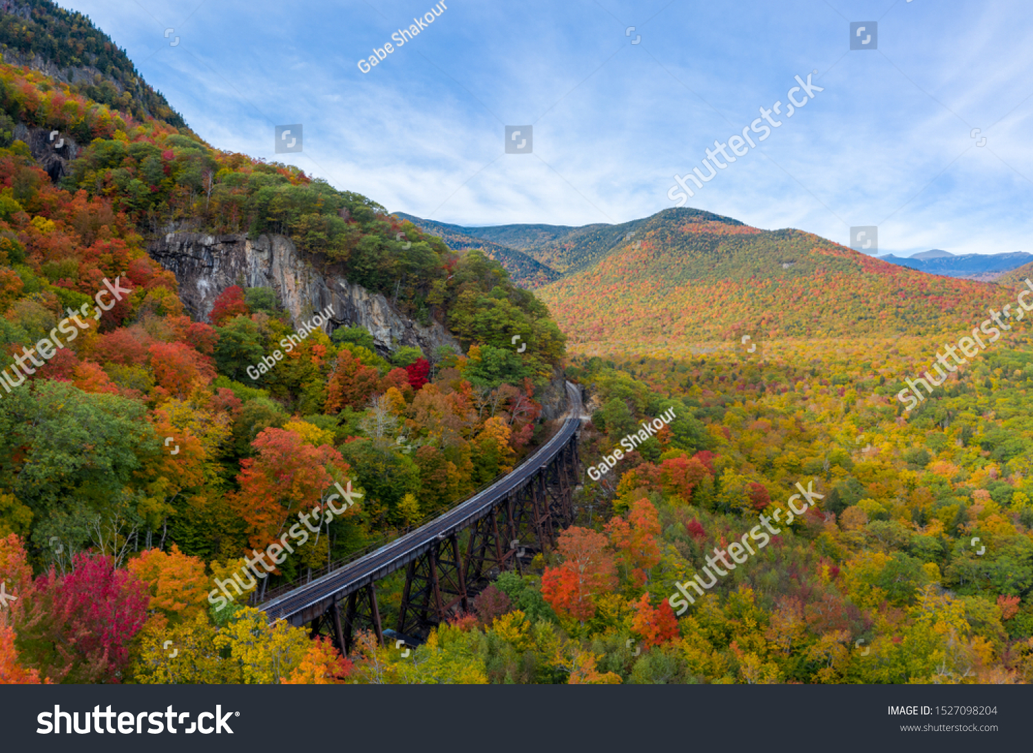 Brightly colored fall foliage during a sunny fall day. Aerial drone shot red, yellow and orange fall leaves with train tracks curving around mountain. #1527098204