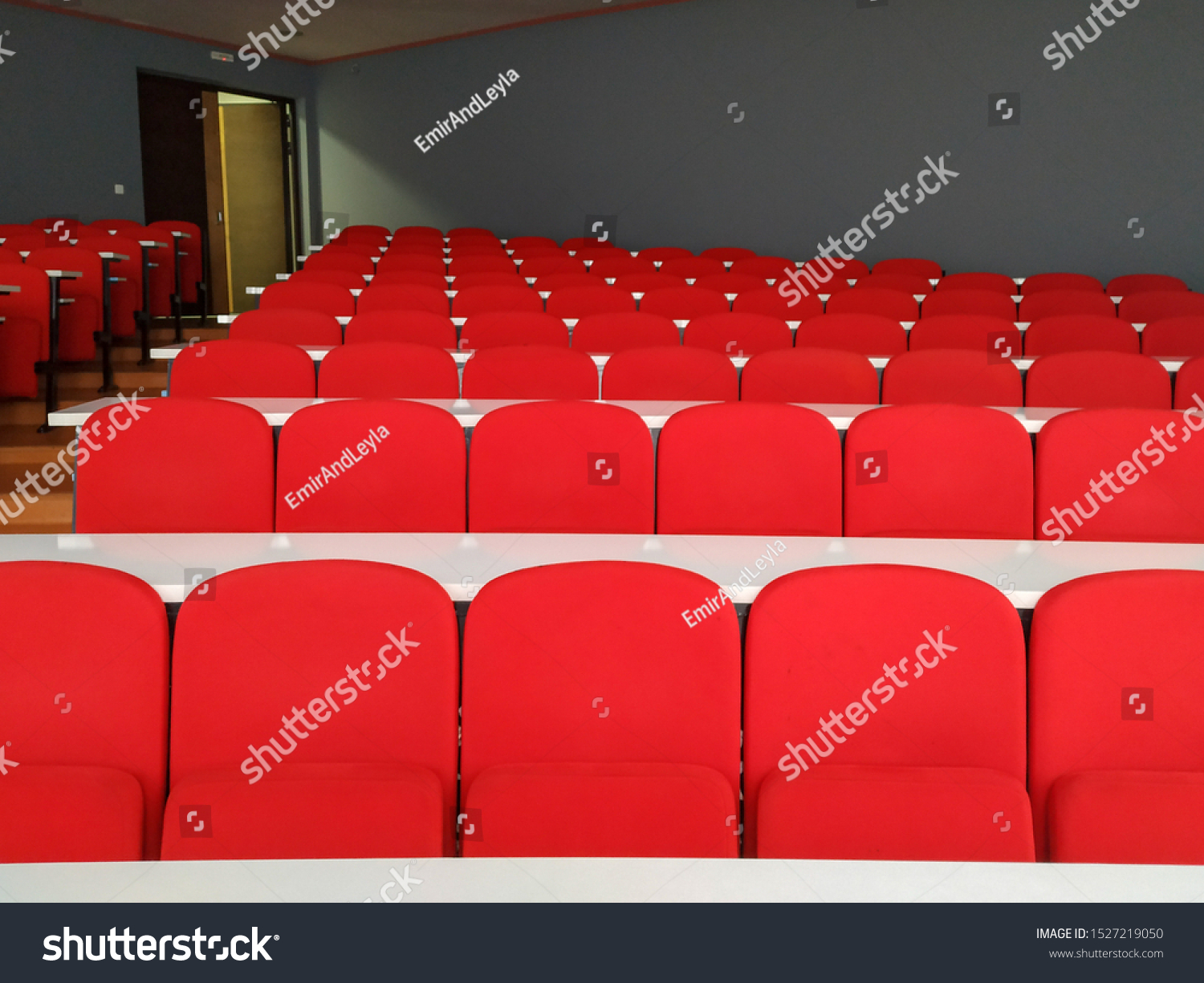Amphitheater big lecture room with red chairs and white tables for students lined in rows for university or conference lectures with doors in background #1527219050