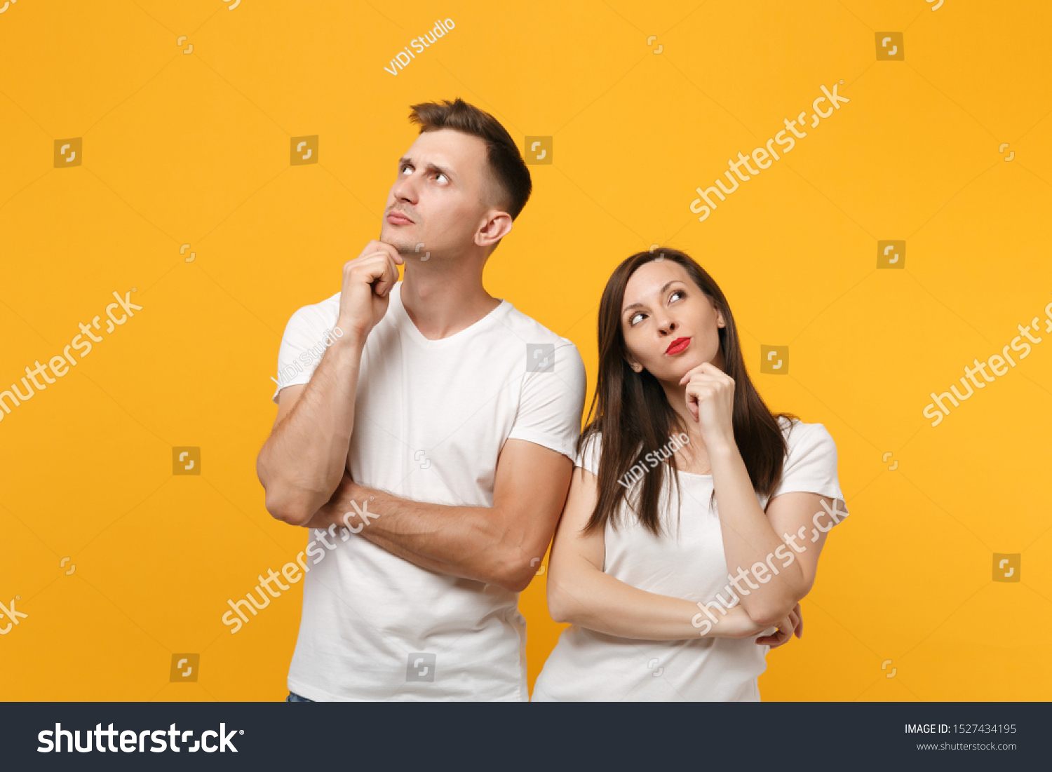 Pensive young couple two friends guy girl in white empty blank t-shirts posing isolated on yellow orange background. People lifestyle concept. Mock up copy space. put hand prop up on chin, looking up #1527434195