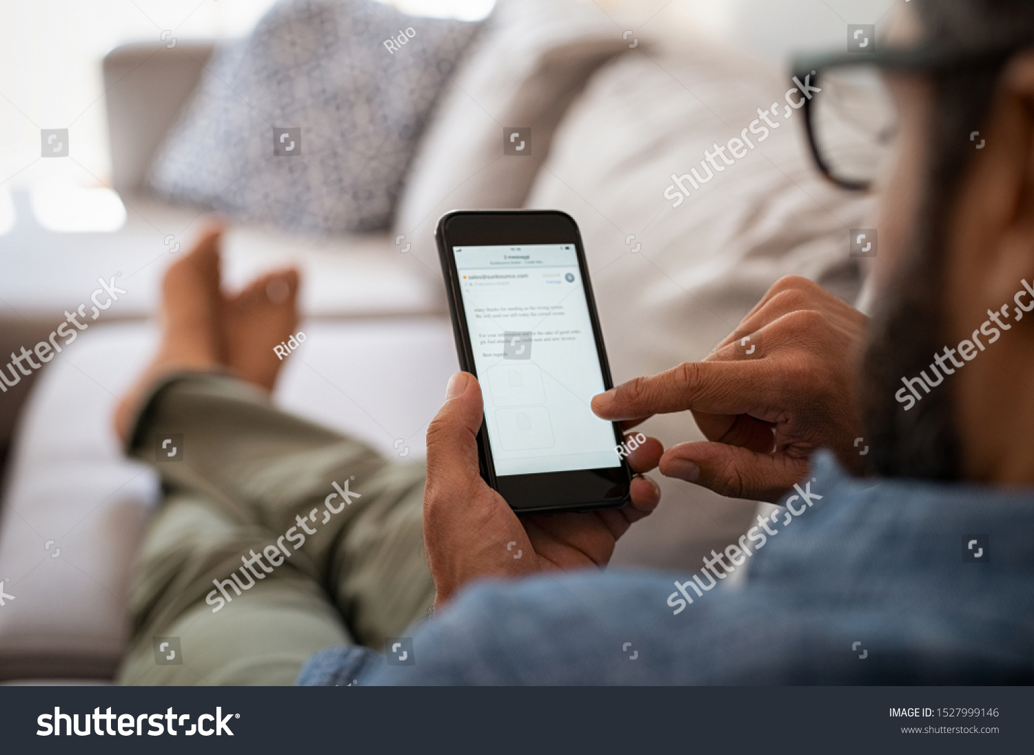 Closeup of a man hand holding cellphone with internet browser on screen. Man with spectacles relaxing sitting on couch. Closeup of mature latin man using smartphone to checking email at home. #1527999146