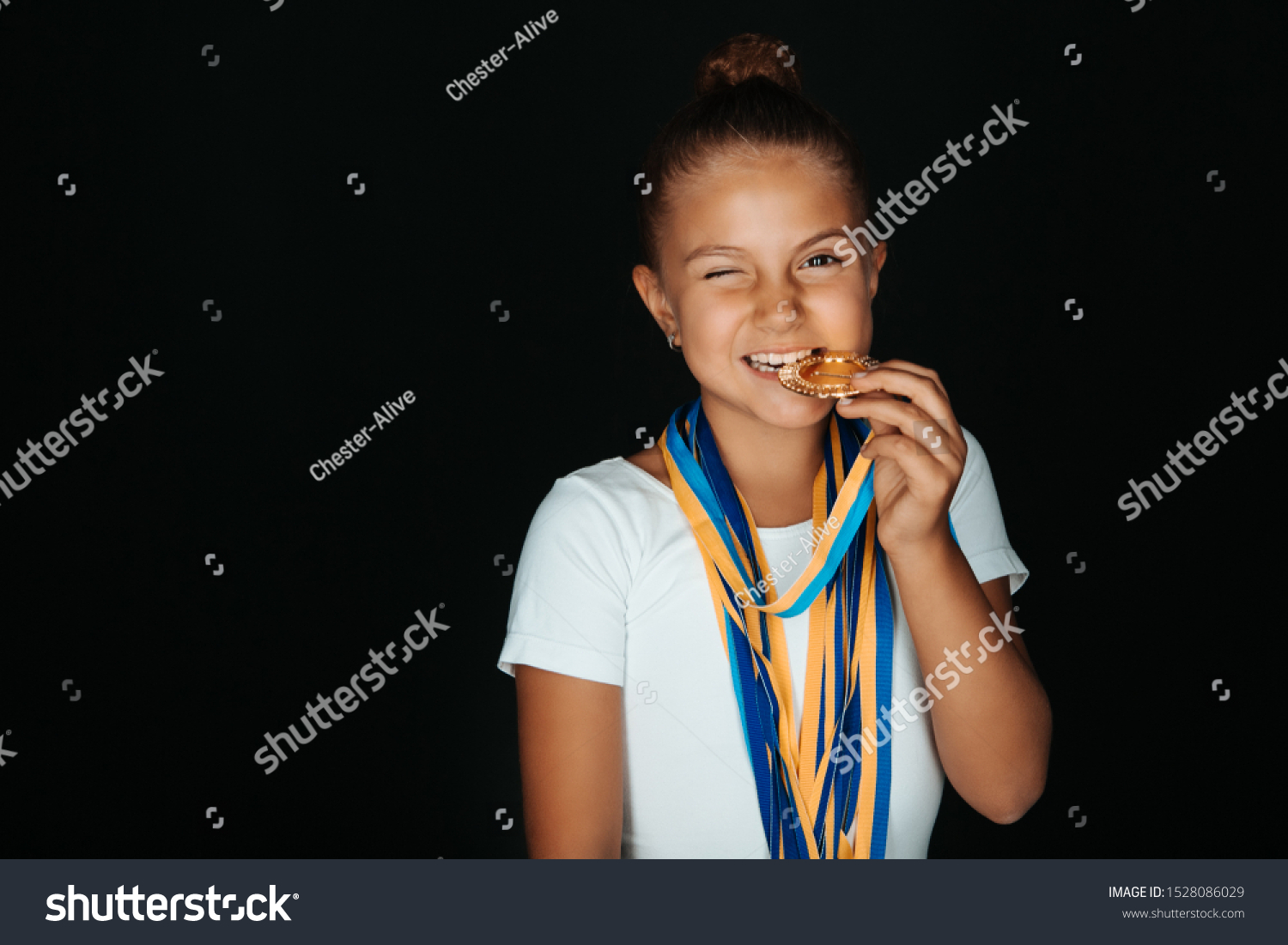 Portrait of little gymnast girl in white bodysuit with medals on her neck biting a medal and winking isolated on black background #1528086029