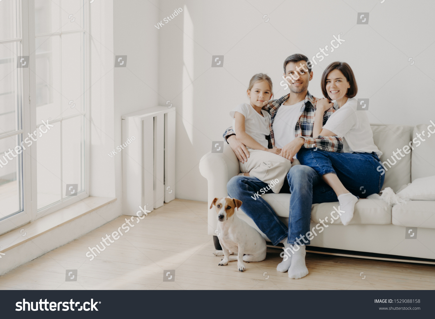 Family, togetherness and relationnship concept. Happy man embraces daughter and wife, sit on comfortable white sofa in empty room, their pet sits on floor, make family portrait for long memory #1529088158