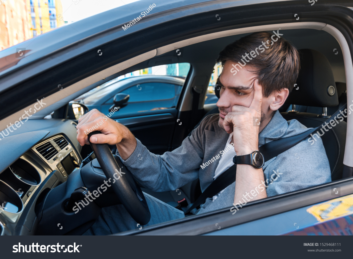 Annoyed tired young man is riding driving car. Businessman is late for meeting. Driver brunette in grey suit stuck in traffic jam. Stressful situations on roads and fast rhythm in modern city. #1529468111