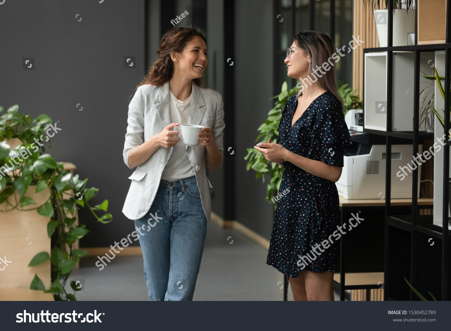 Asian and Caucasian ethnicity women colleagues met in office hall chatting enjoy friendly warm conversation, multi-ethnic mates having informal talk drink tea or coffee take break distracted from work #1530452789
