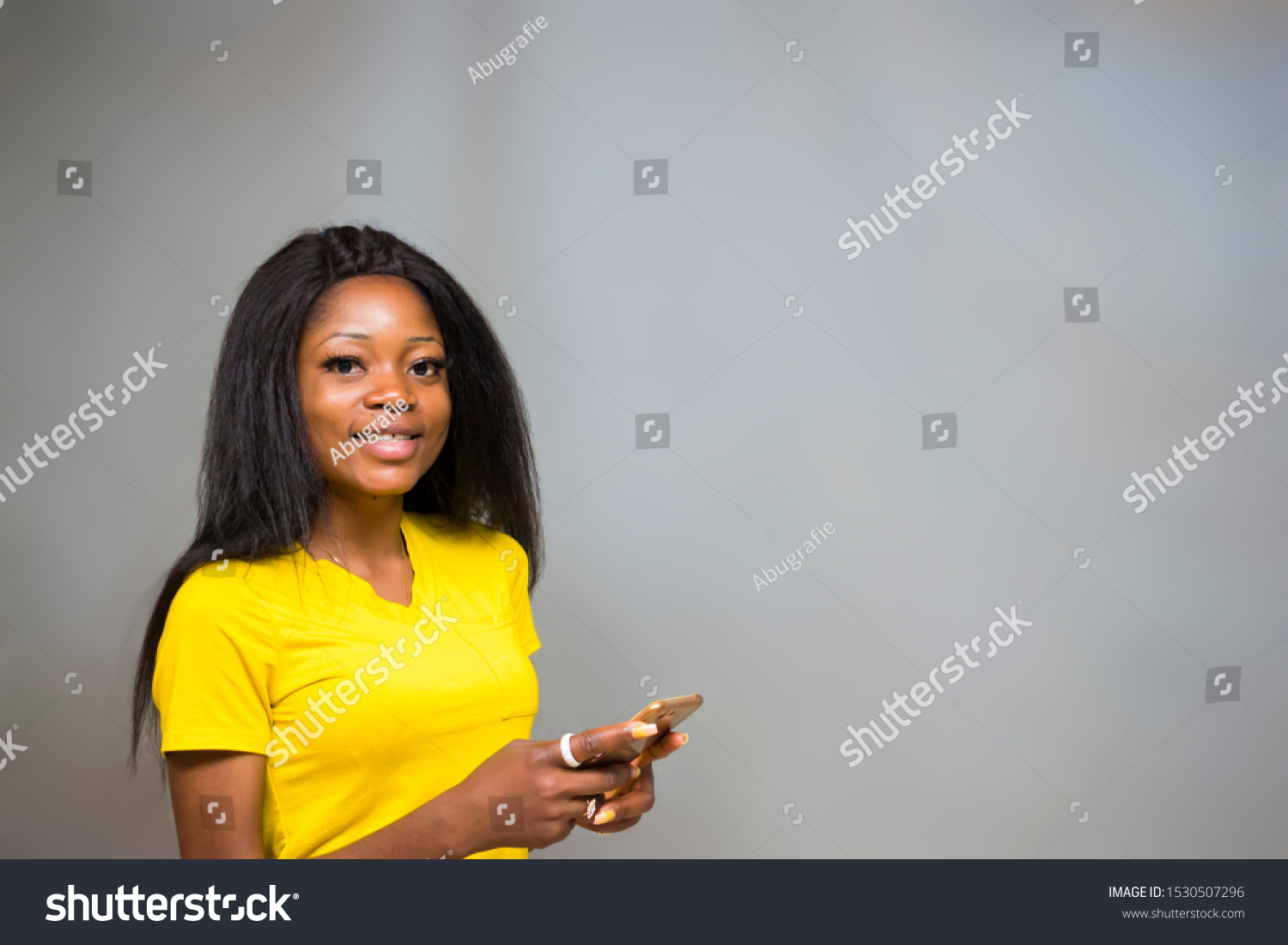 satisfied Young African hipster girl,types text message on cell phone, enjoys online communication, types feedback, wears yellow shirt, isolated on Gray studio wall. Technology concept #1530507296