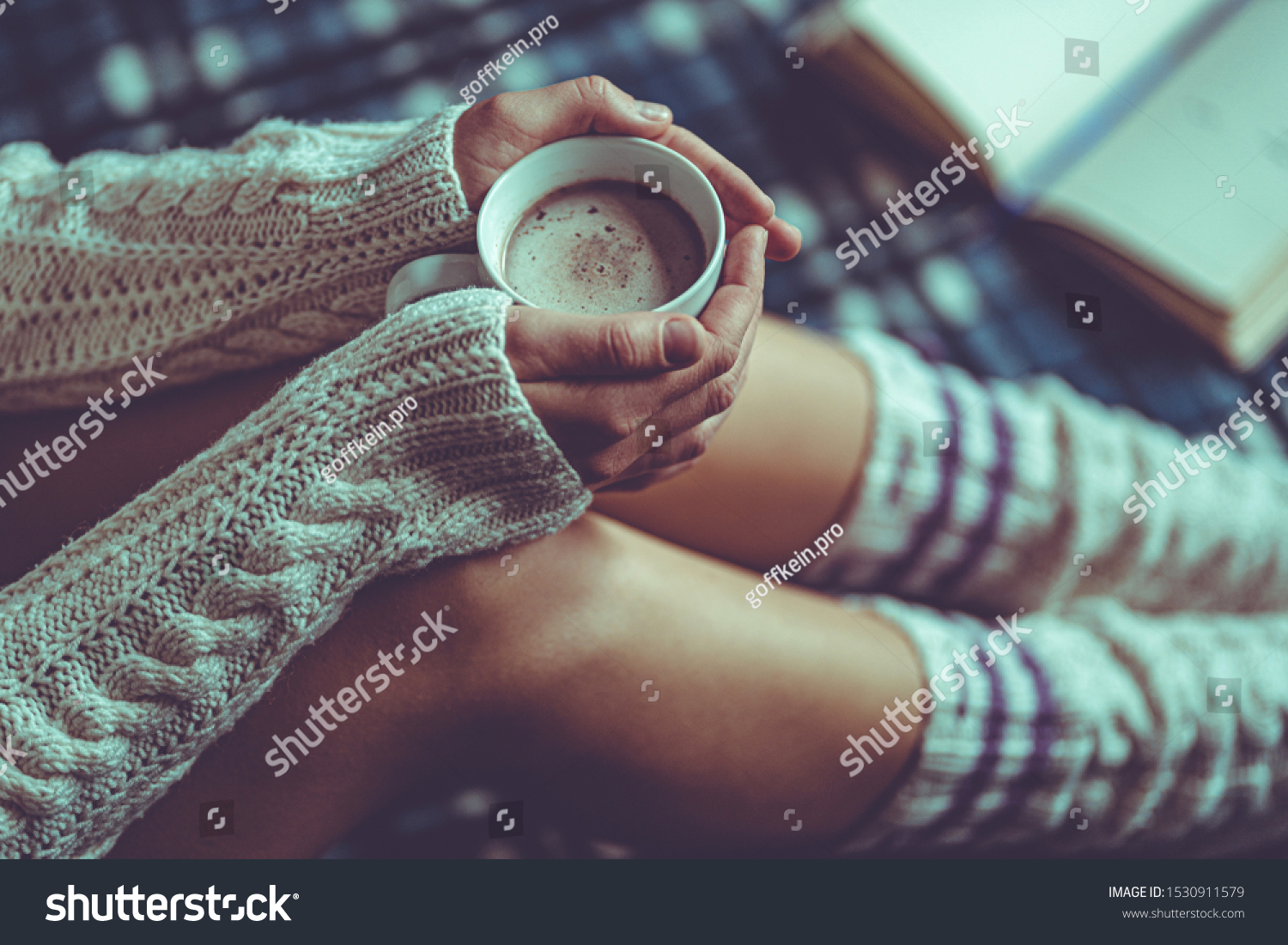 Comforting cozy woman in knitted winter socks and white warm sweater sitting on a plaid blanket and warming up with a cup of hot cocoa at home in winter time. Cozy time and winter drinks  #1530911579