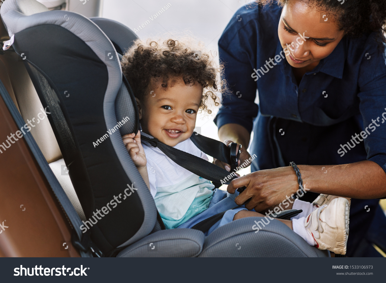 Side view of a happy little boy looking at camera while his mother buckling him in a car seat #1533106973