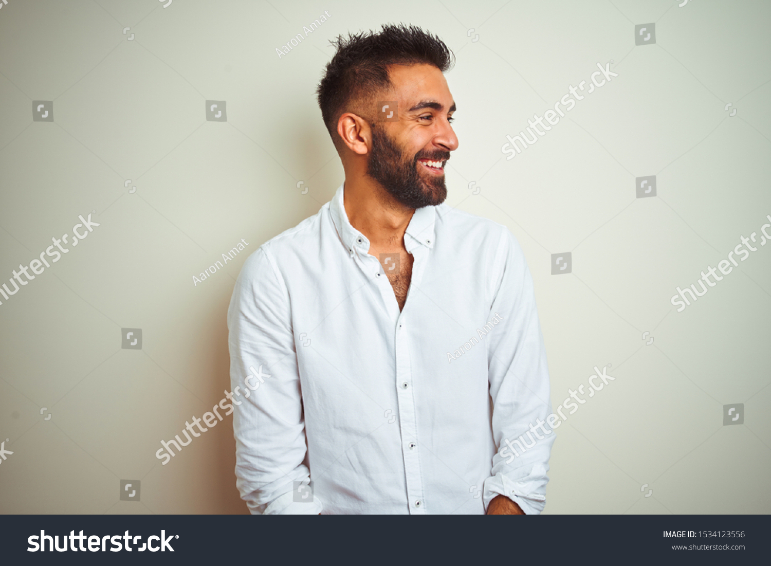Young indian man wearing elegant shirt standing over isolated white background looking away to side with smile on face, natural expression. Laughing confident. #1534123556