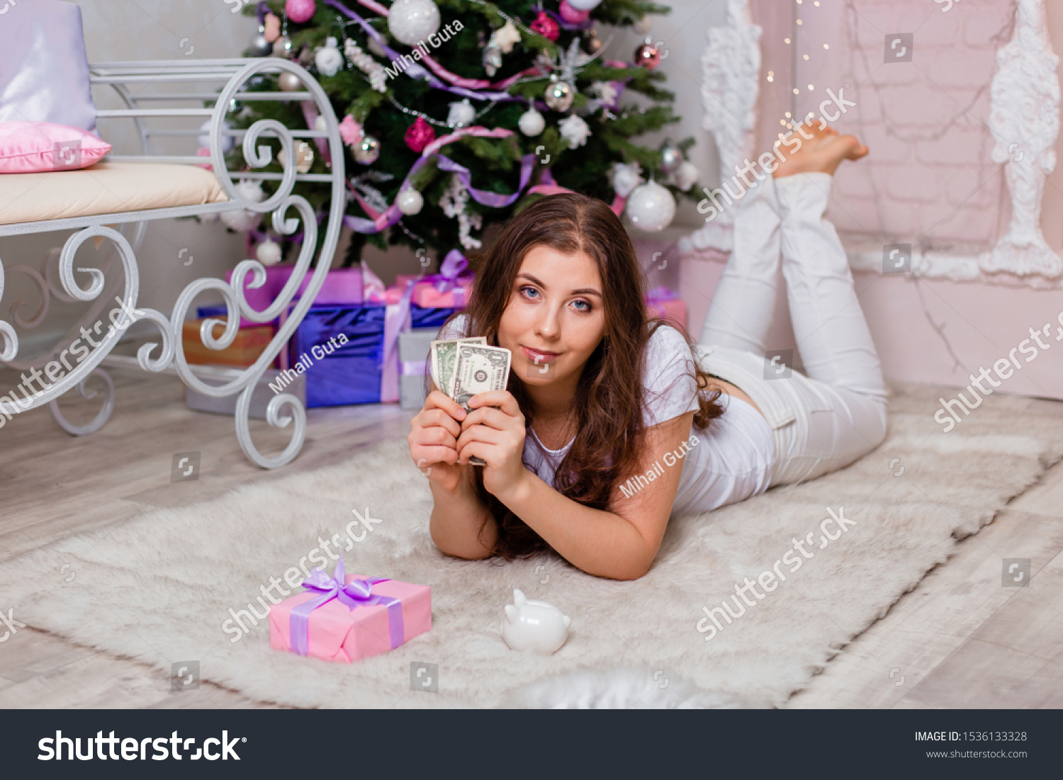 An attractive girl in a white T-shirt lies on her stomach on a fluffy fur rug with dollars in her hands next to a gift and a piggy bank near the fireplace and Christmas tree. #1536133328