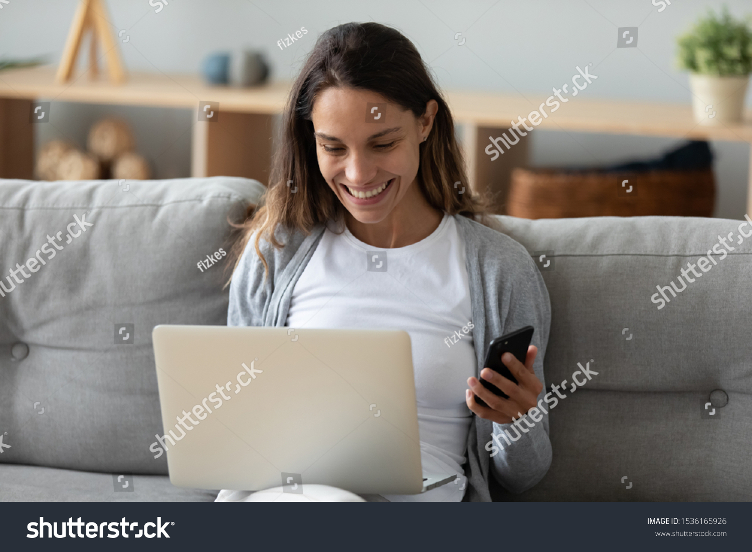 Head shot smiling young mixed race lady sitting on sofa at home with computer, shopping in internet, making purchases in online store, entering payment confirmation code from mobile phone message. #1536165926