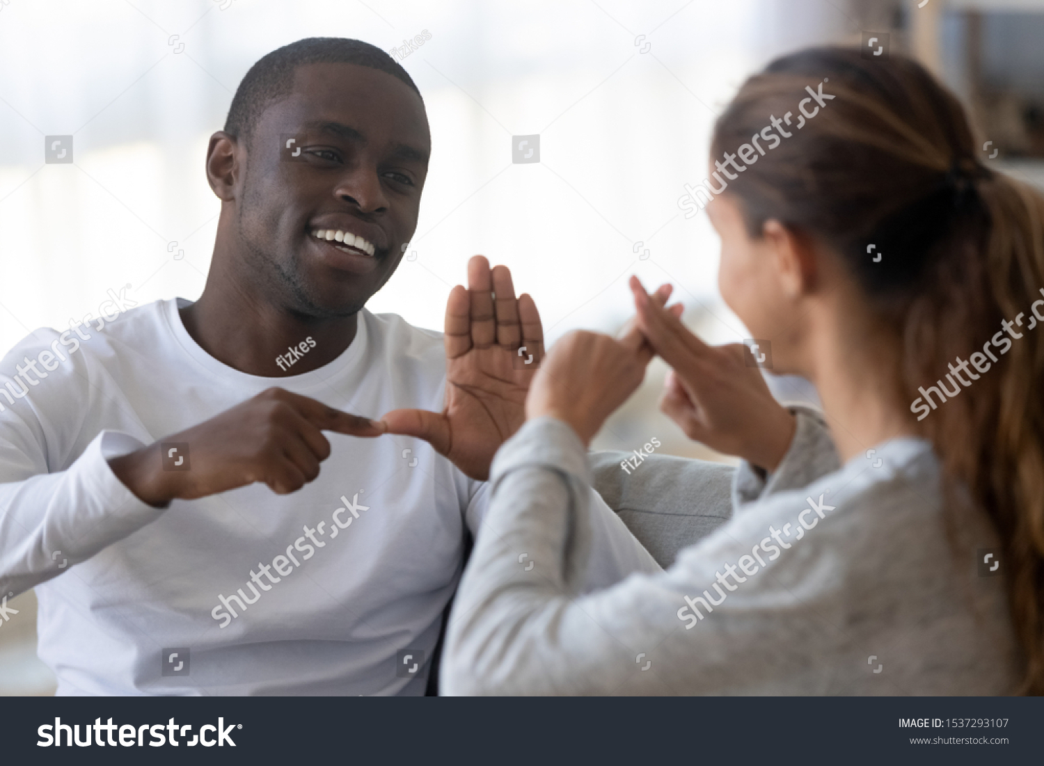 Smiling african American man sit on couch show hand gestures talking with female friend at home, international disabled hearing impaired couple or spouses use sign language communicating #1537293107