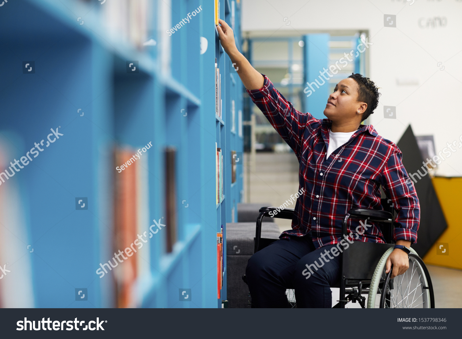 Portrait of disabled student in wheelchair choosing books while studying in college library, copy space #1537798346