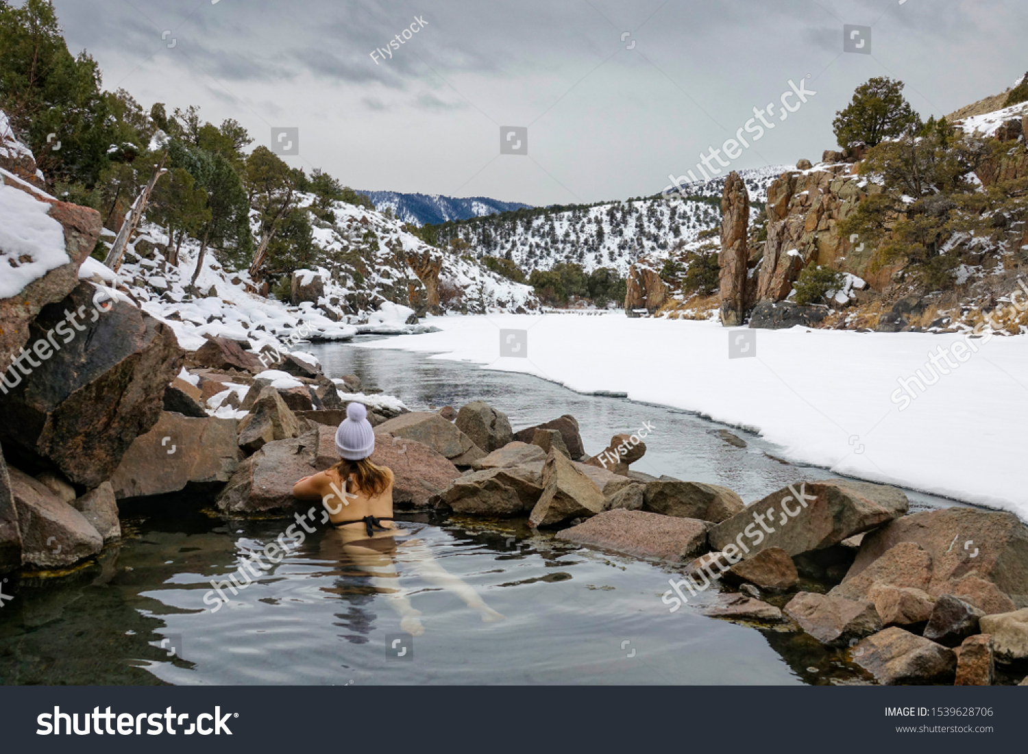 CLOSE UP: Young woman hiking in Colorado relaxes in the Radium Hot springs. Girl in a black bikini and white winter cap lies in the therapeutic water of ahot pond and observes the snowy nature. #1539628706