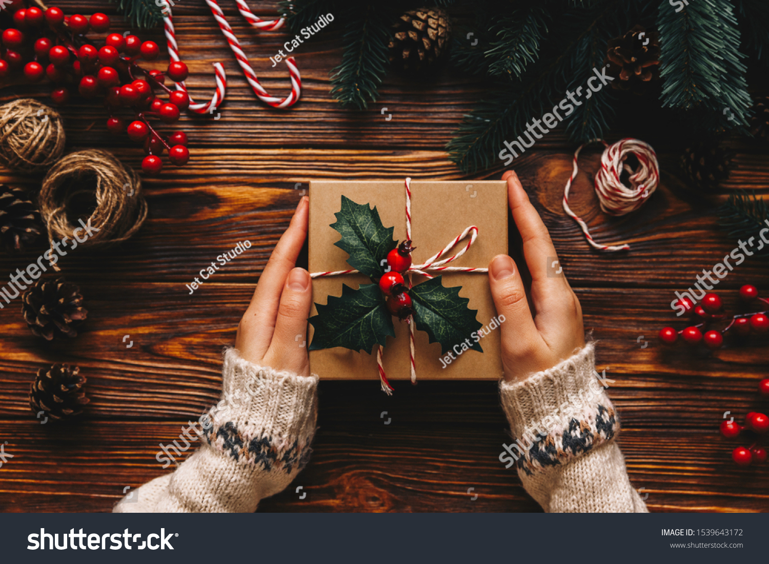 Christmas gift. Congratulations.Top wiew of woman holding traditional decorated gift box. Wooden table with cane,fir branches,fir cone, and berries #1539643172