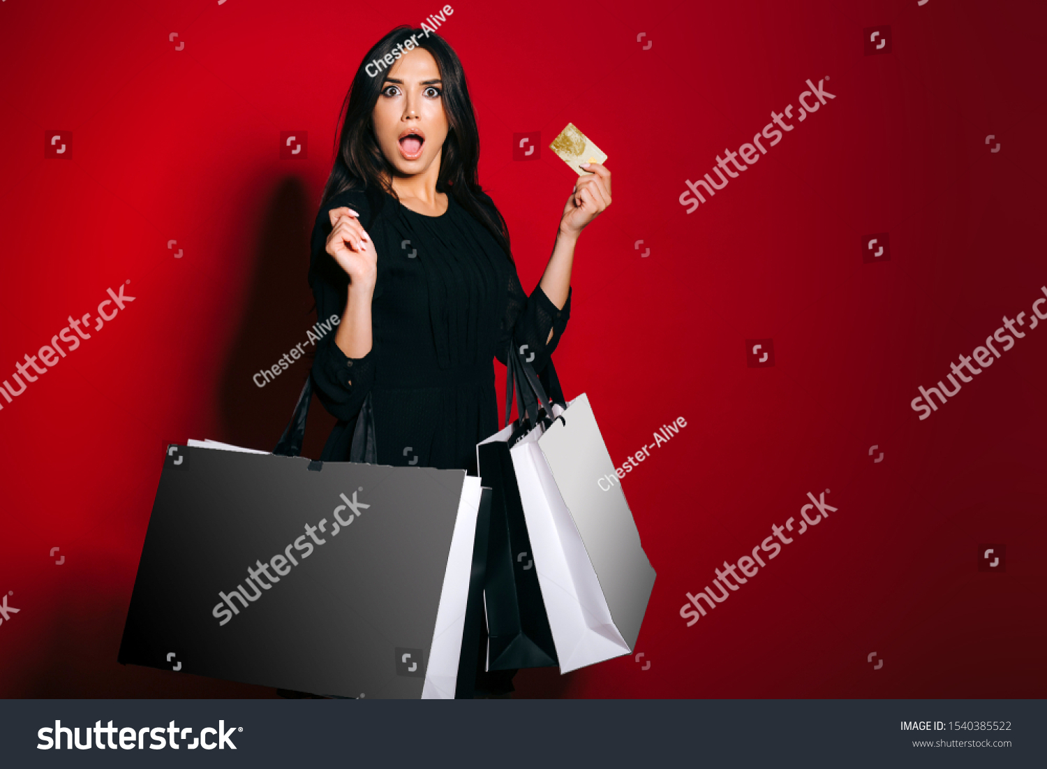 Surprised glamourous woman carrying shopping bags holding a gold credit card on dark red background. #1540385522