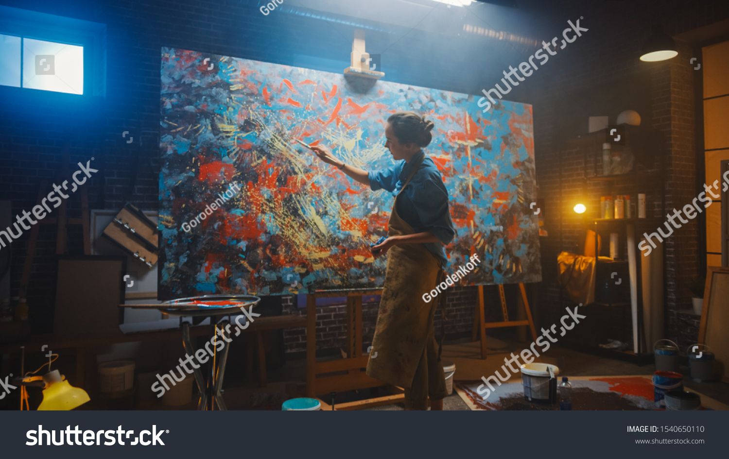 Talented Female Artist Working on a Modern Abstract Oil Painting, Gesturing with Broad Strokes Using Paint Brush. Dark Creative Studio Large Picture Stands on Easel Illuminated, Tools Everywhere #1540650110