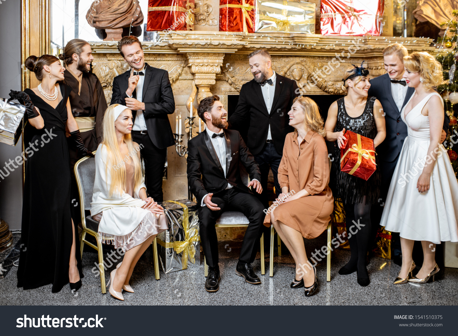 Staged group photo of an elegant well-dressed people near the beautifully decorated fireplace with christmas tree and presents during a New Year celebration #1541510375