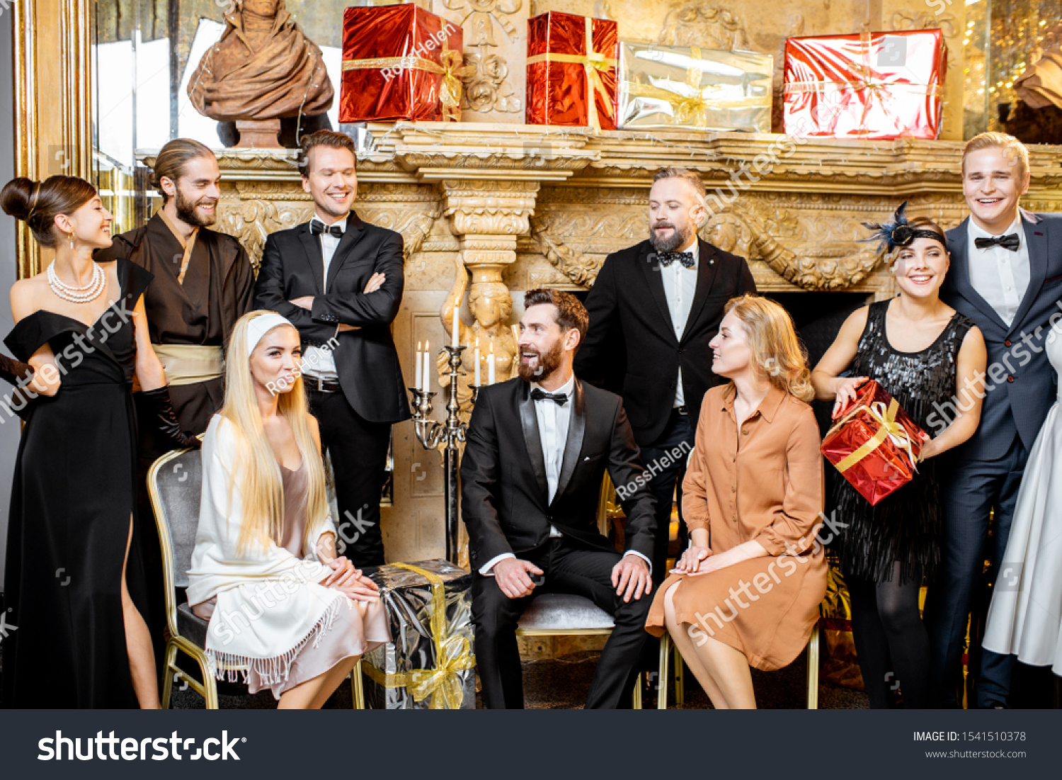 Staged group photo of an elegant well-dressed people near the beautifully decorated fireplace with christmas tree and presents during a New Year celebration #1541510378