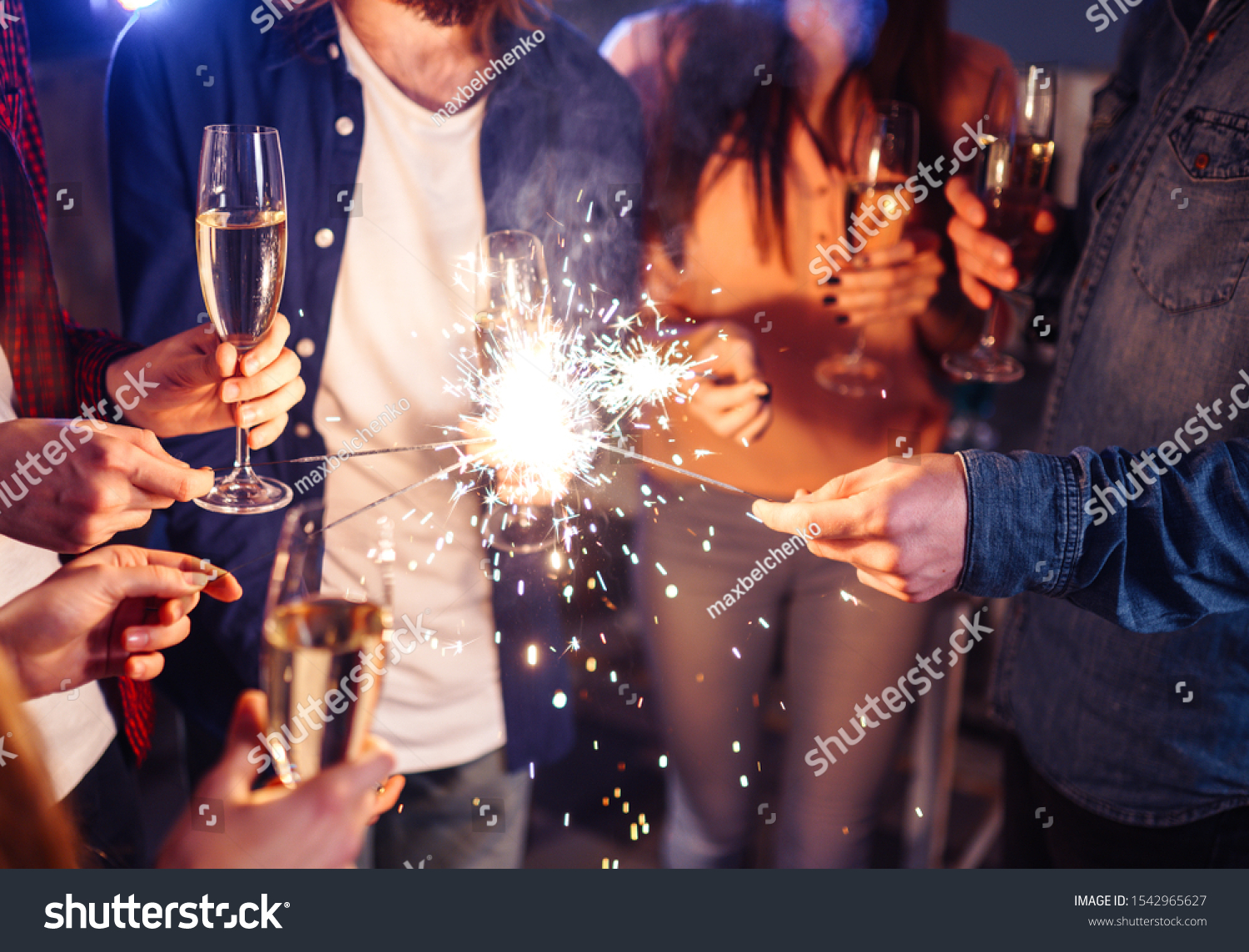 Group of happy people holding sparklers at party and smiling. Young people celebrating New Year together. Friends lit sparklers. Friends enjoying with sparklers in evening. Blur Background. #1542965627