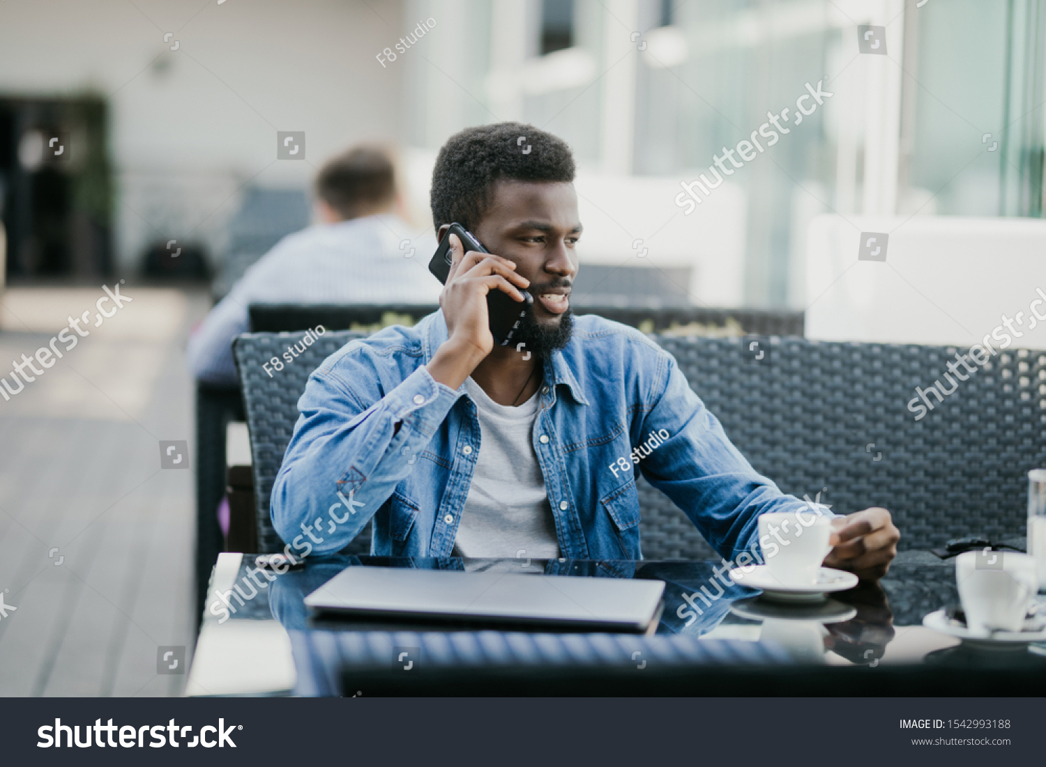 Smiling young african man drinking coffee and talking on mobile phone while sitting in cafe, view through the window #1542993188