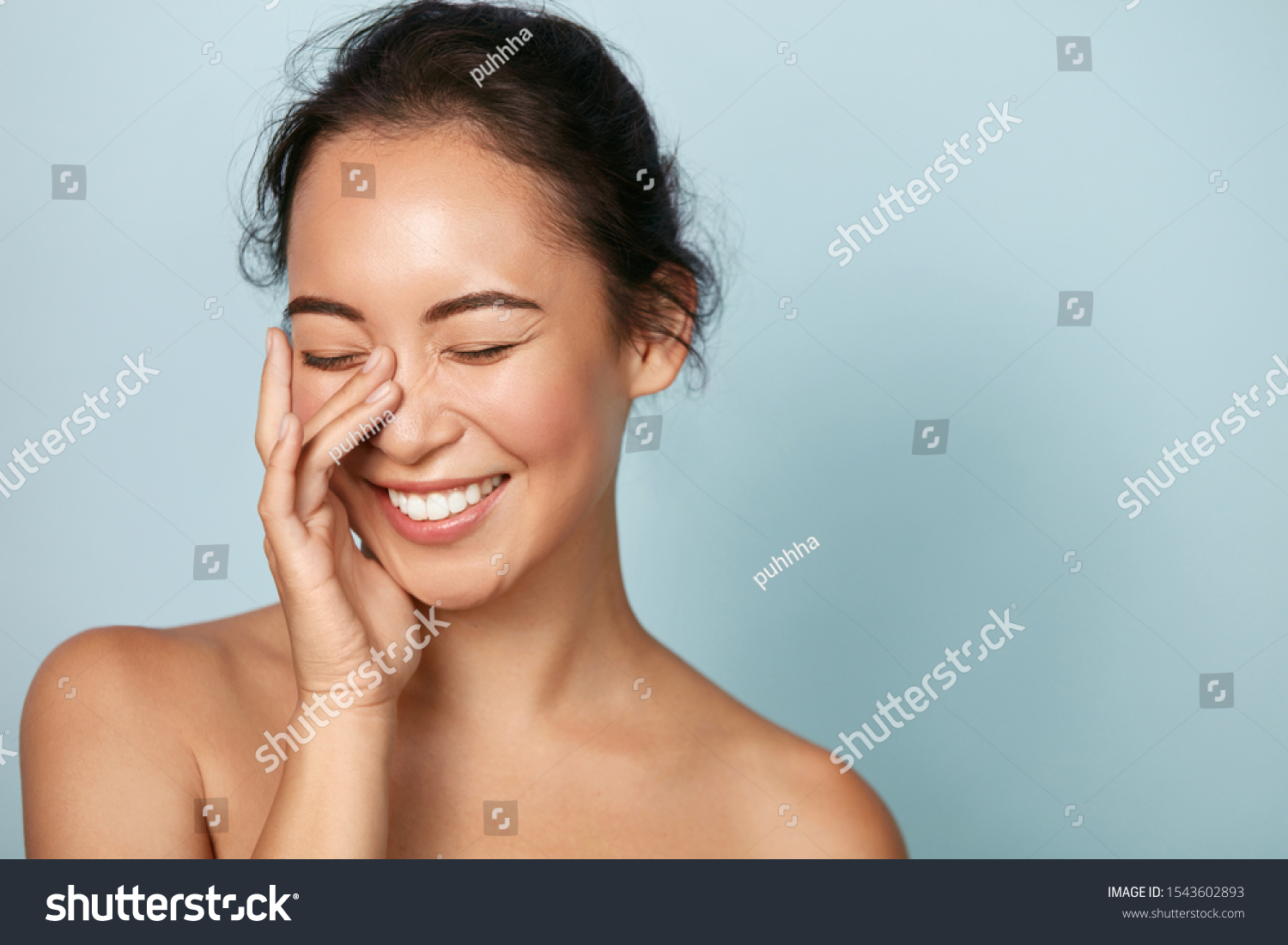 Beauty face. Smiling asian woman touching healthy skin portrait. Beautiful happy girl model with fresh glowing hydrated facial skin and natural makeup on blue background at studio. Skin care concept