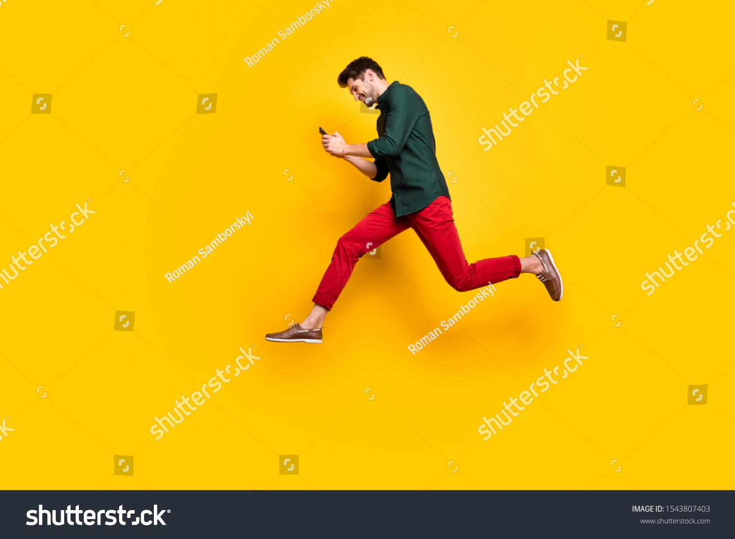Full size profile side photo of positive cheerful brunet hair use his cellphone search find discounts jump run feel  content wear green shirt red pants trousers isolated over yellow color background #1543807403