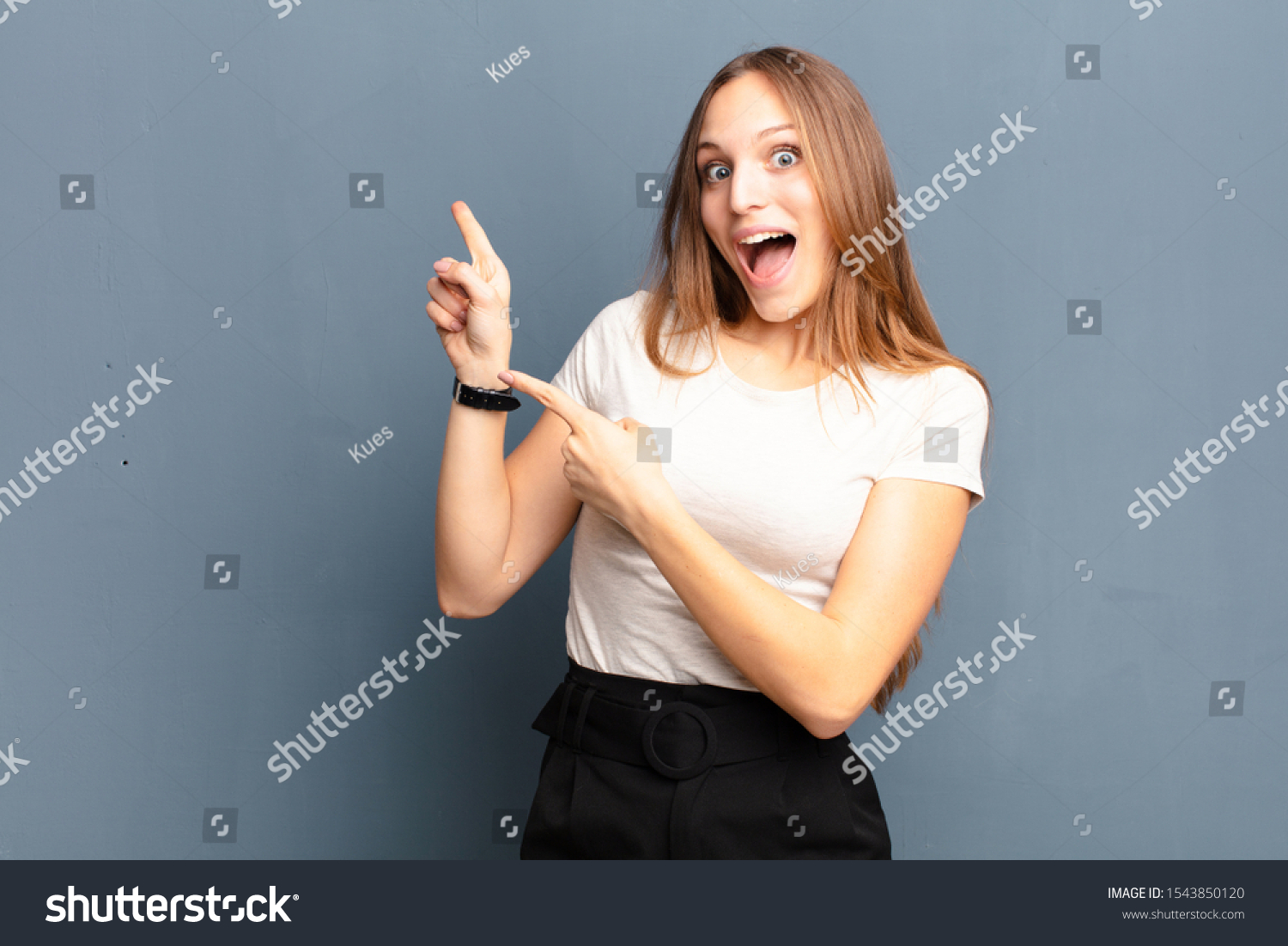 young pretty woman feeling joyful and surprised, smiling with a shocked expression and pointing to the side against gray background #1543850120