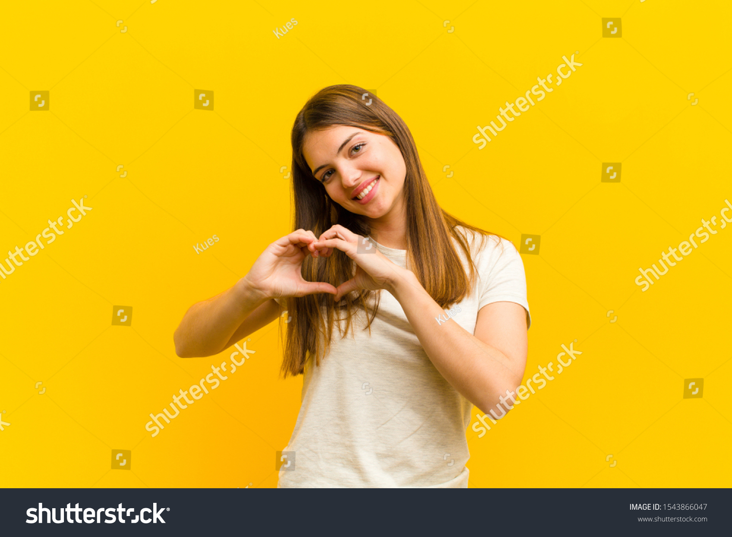 young pretty woman smiling and feeling happy, cute, romantic and in love, making heart shape with both hands against orange background #1543866047