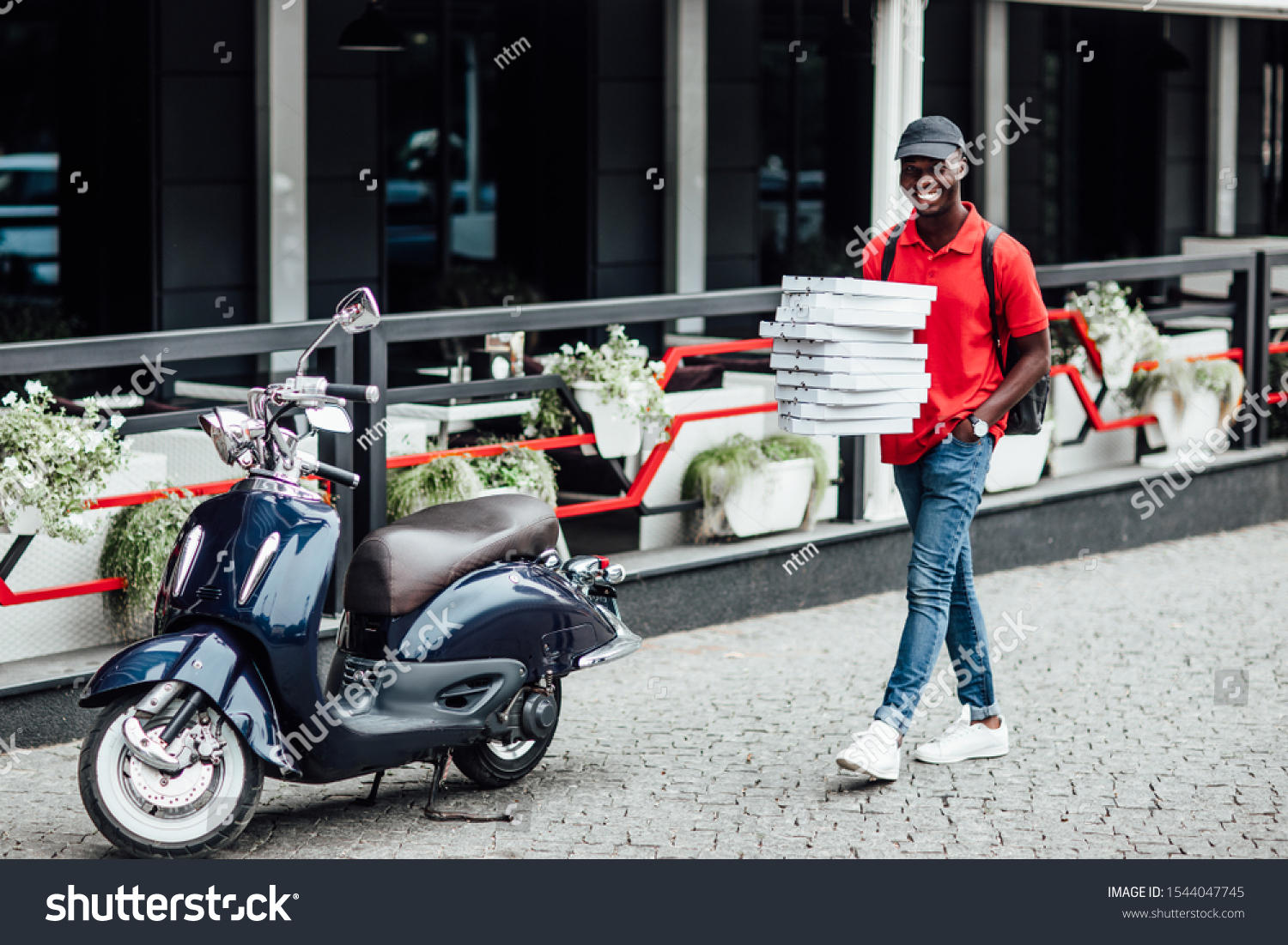 Busy deliveryman being in hurry, carries cardboard boxes with pizza, delivers to customers, poses on blue scooter, wears helmet, white t shirt and sportshoes, spreads legs, has happy expression. #1544047745