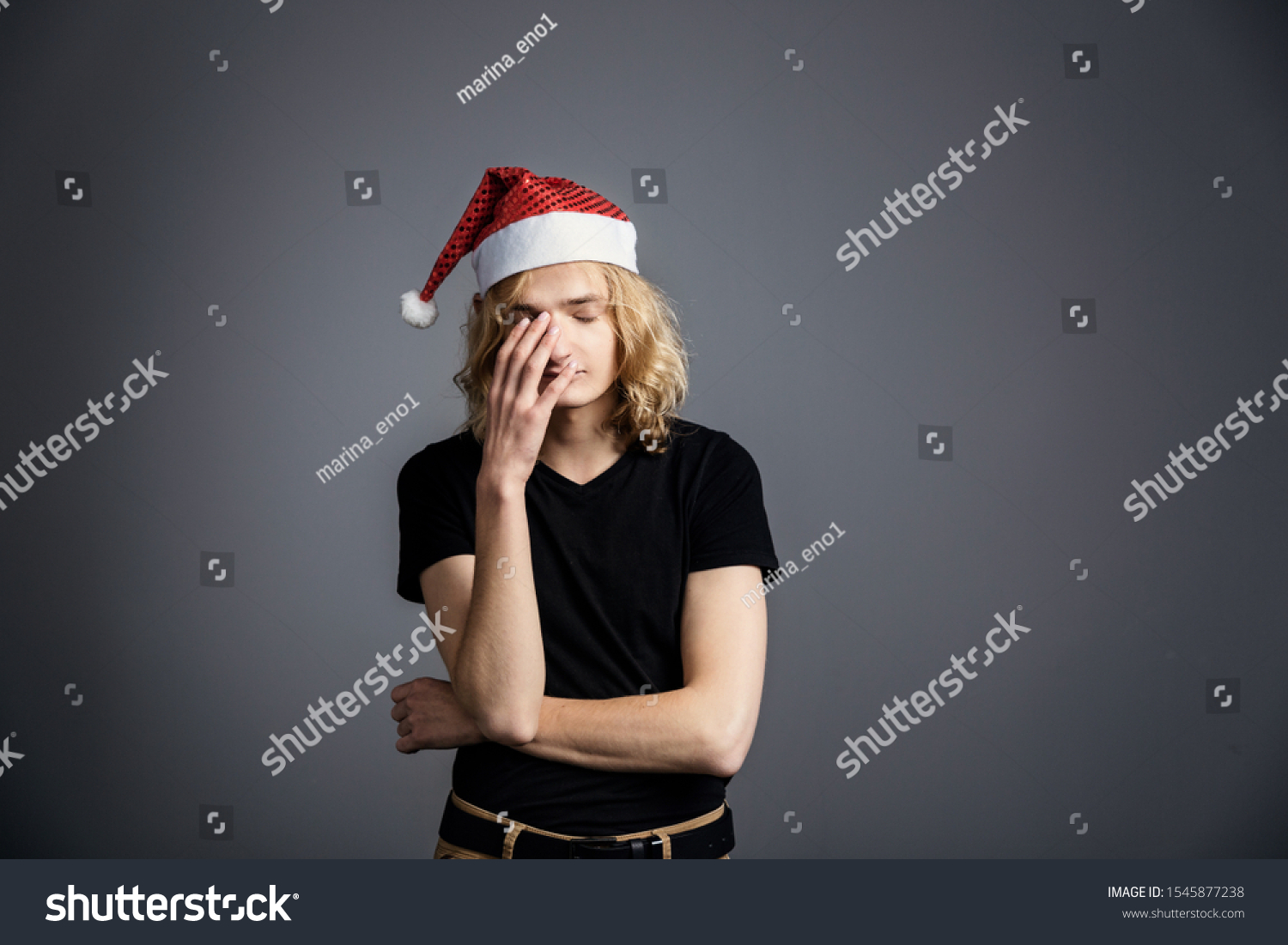 Young guy an attractive guy with long blonde hair in the form of a Christmas elf in a Santa Claus hat is standing with a displeased skeptical face on a gray background. covered his face with his hand  #1545877238