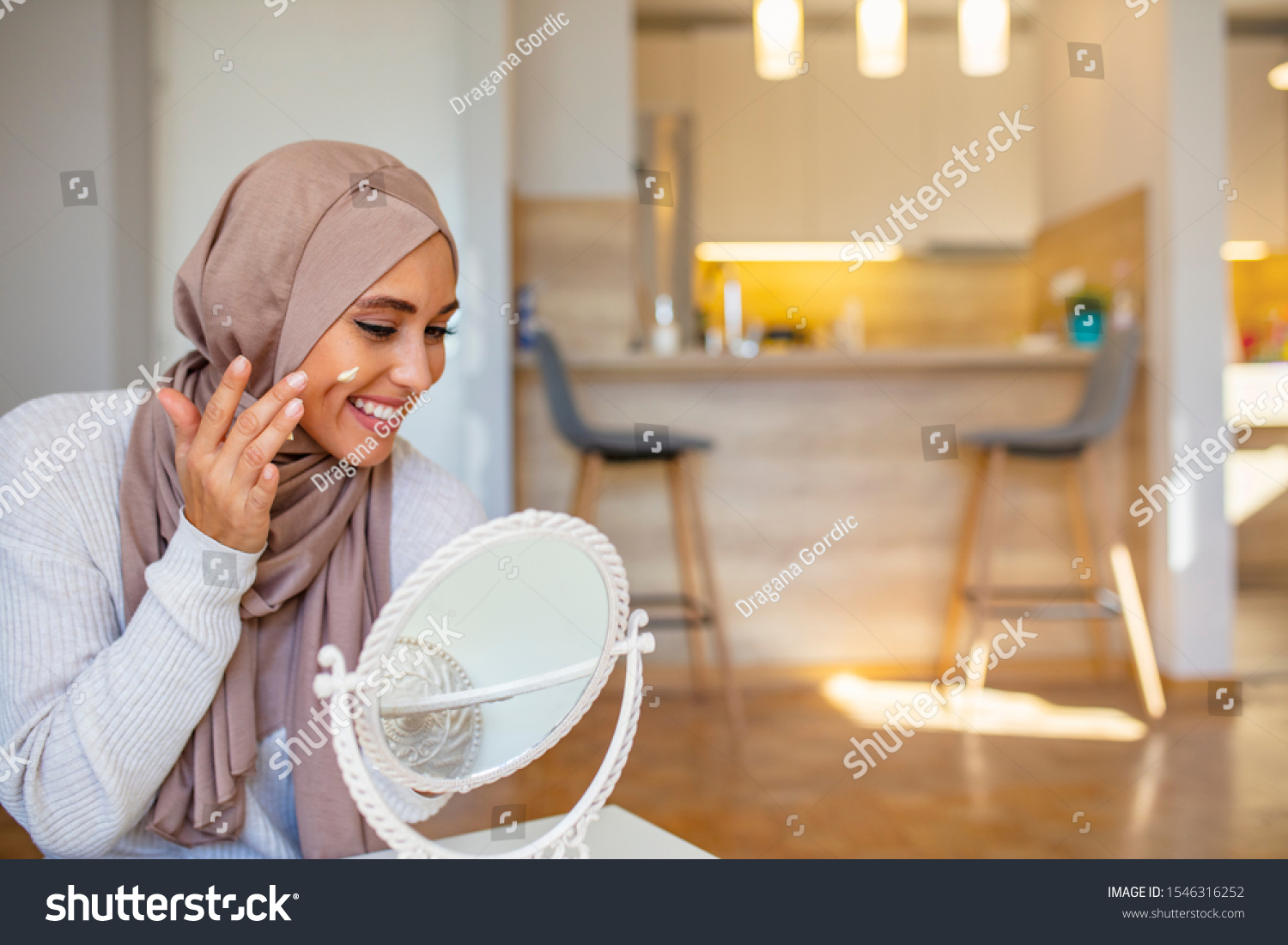 Muslim woman spreading cream over her face while looking in the mirror. Beauty treatment. Female putting on moisturizer on her facial. Smiling Muslim woman applying cream to face and looking to mirror #1546316252