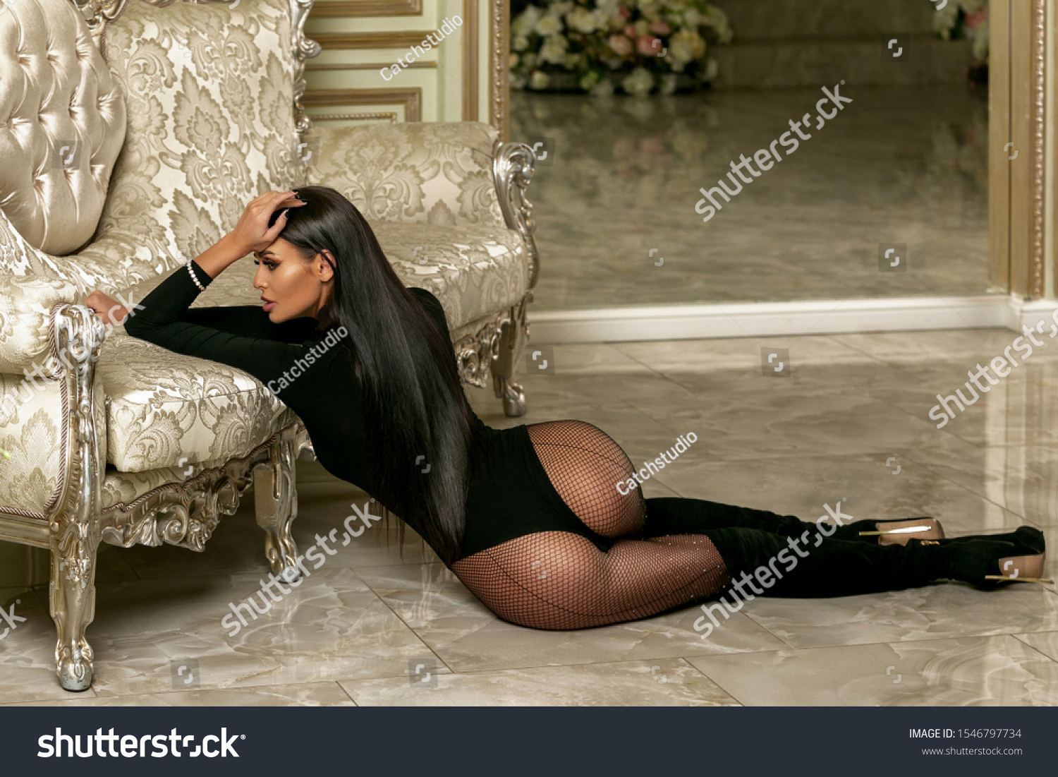 Beautiful young girl in a black bodysuit and boots in the living room. Attractive brunette model with long legs sits on the floor near the sofa and poses. Successful life theme. Horizontal photo. #1546797734