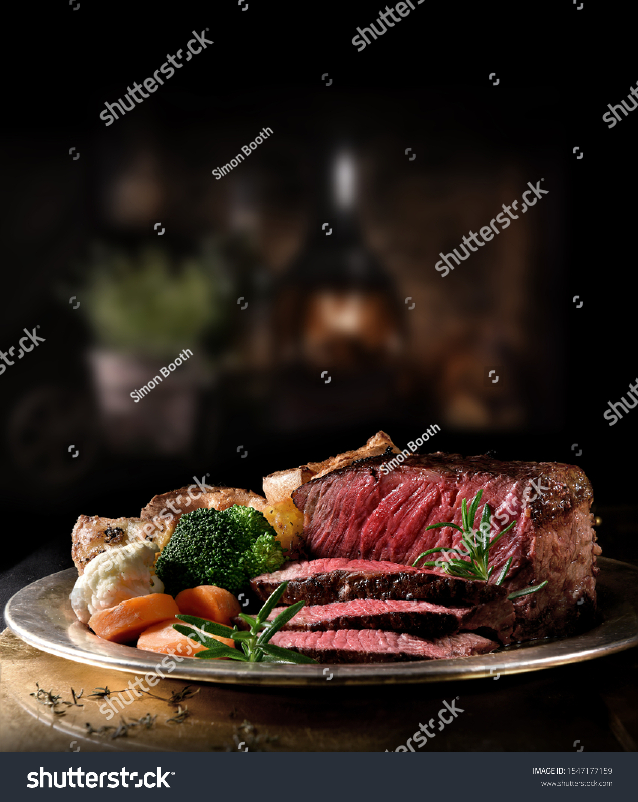 Succulent side of rare roast beef with seasonal vegetables, Yorkshire Puddings and roast potatoes with Rosemary garnish shot in a rustic setting with an old fashioned wood burner. Copy space. #1547177159