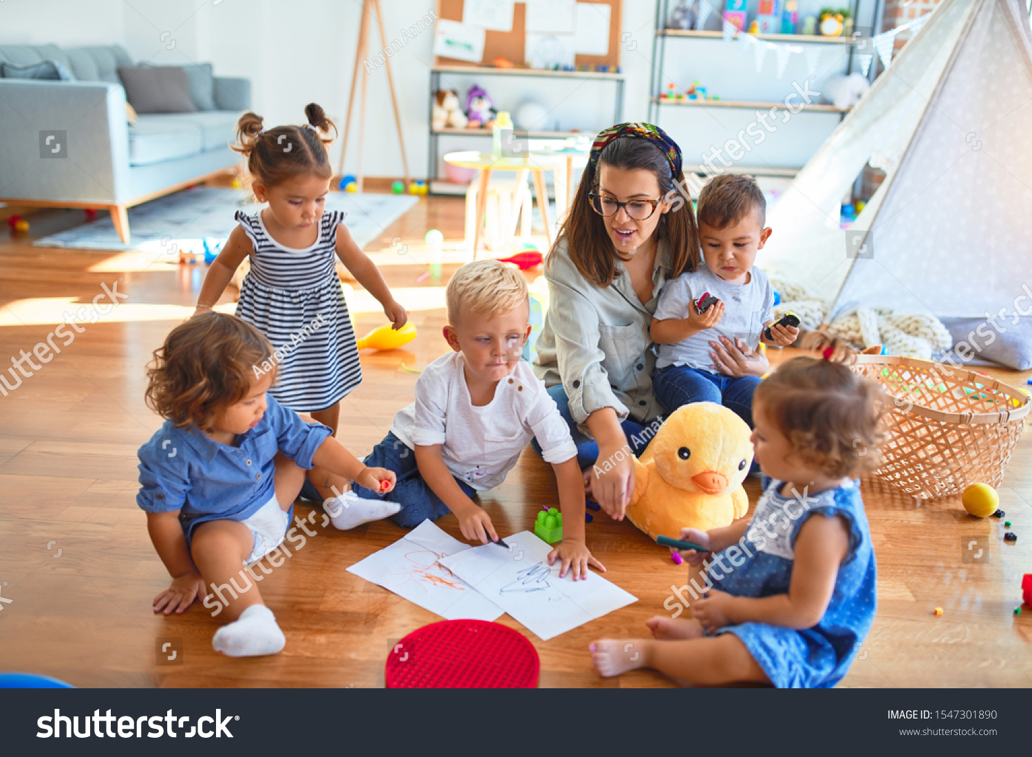 Beautiful teacher and group of toddlers sitting on the floor drawing using paper and pencil around lots of toys at kindergarten #1547301890