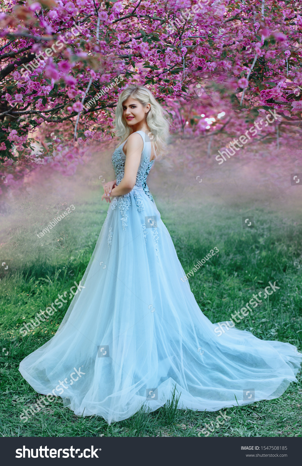An incredible, delicate blonde in a luxurious, delicately blue dress walks in the spring, sakura and cherry orchards are in bloom. Princess with long curly hair. Vanilla color art photo #1547508185