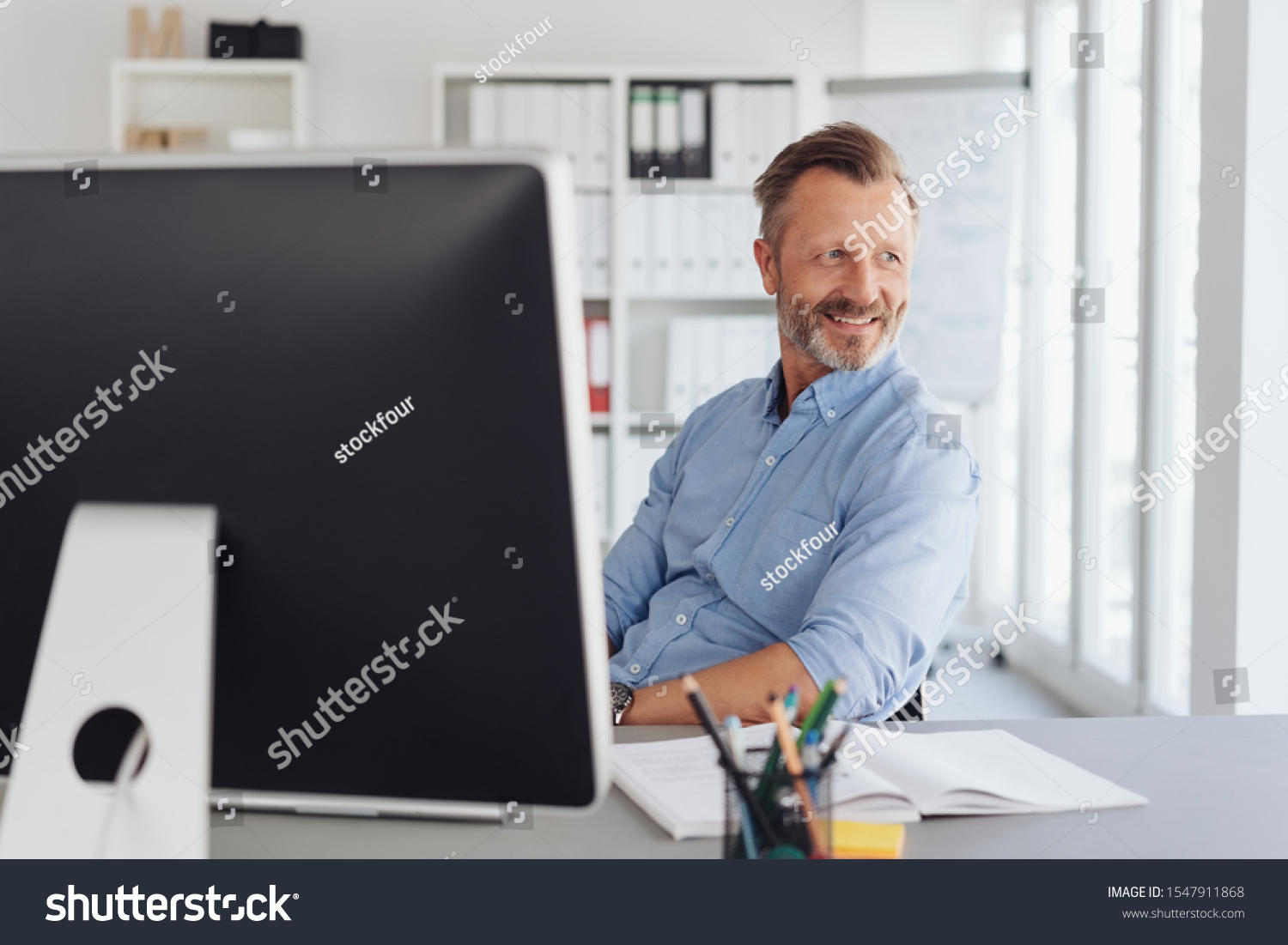 Relaxed businessman watching in amusement as he sits back in his chair looking back through an office window #1547911868