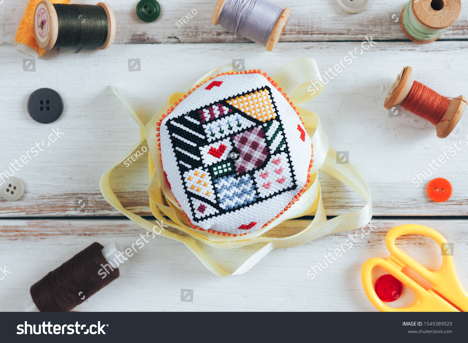 Handmade and needlework. Handmade pin cushion with a simple ornament and a pair of scissors on a white table. Sewing supplies in the background. Top view #1549389929