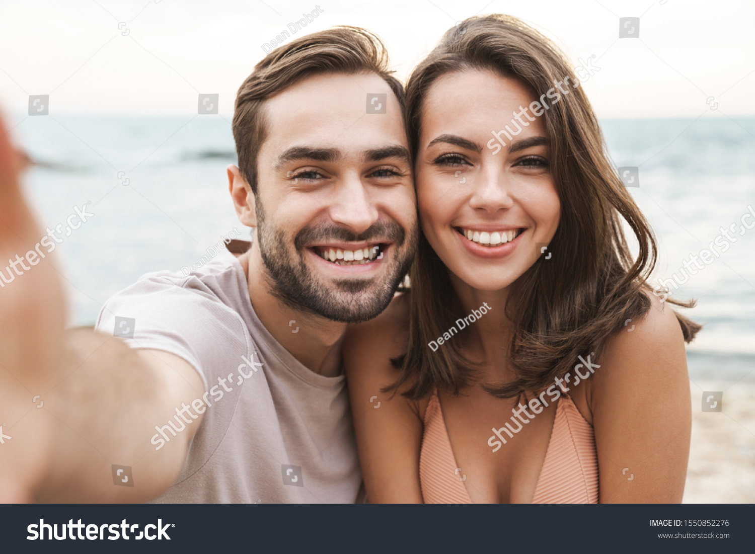 Photo of joyful young couple smiling and taking selfie photo while resting on sunny beach #1550852276