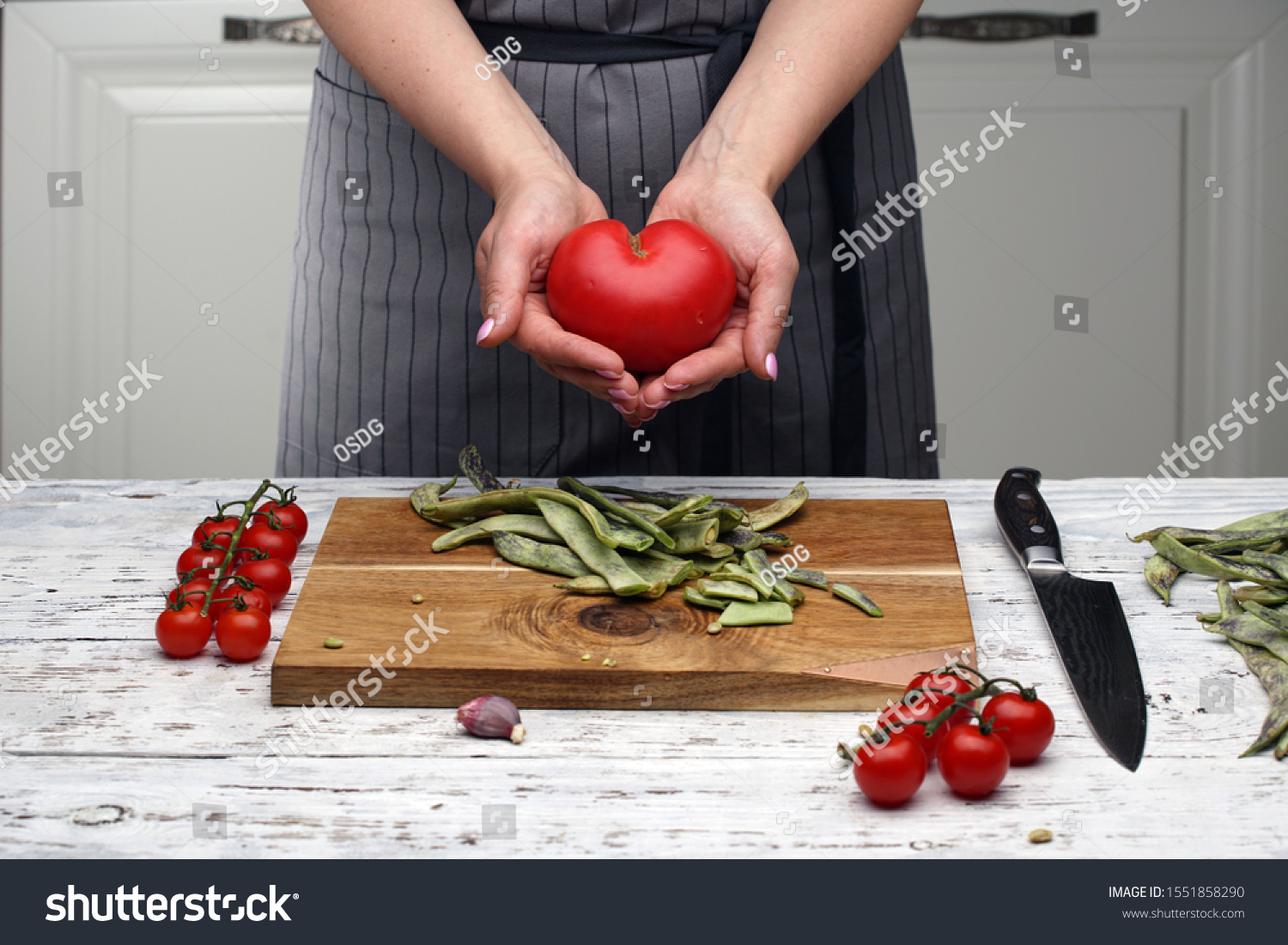 a heart-shaped tomato. a woman's hands hold a heart-shaped tomato against a gray black-striped apron on a white kitchen. cooking healthy food. Love and health concept. cook with love. #1551858290
