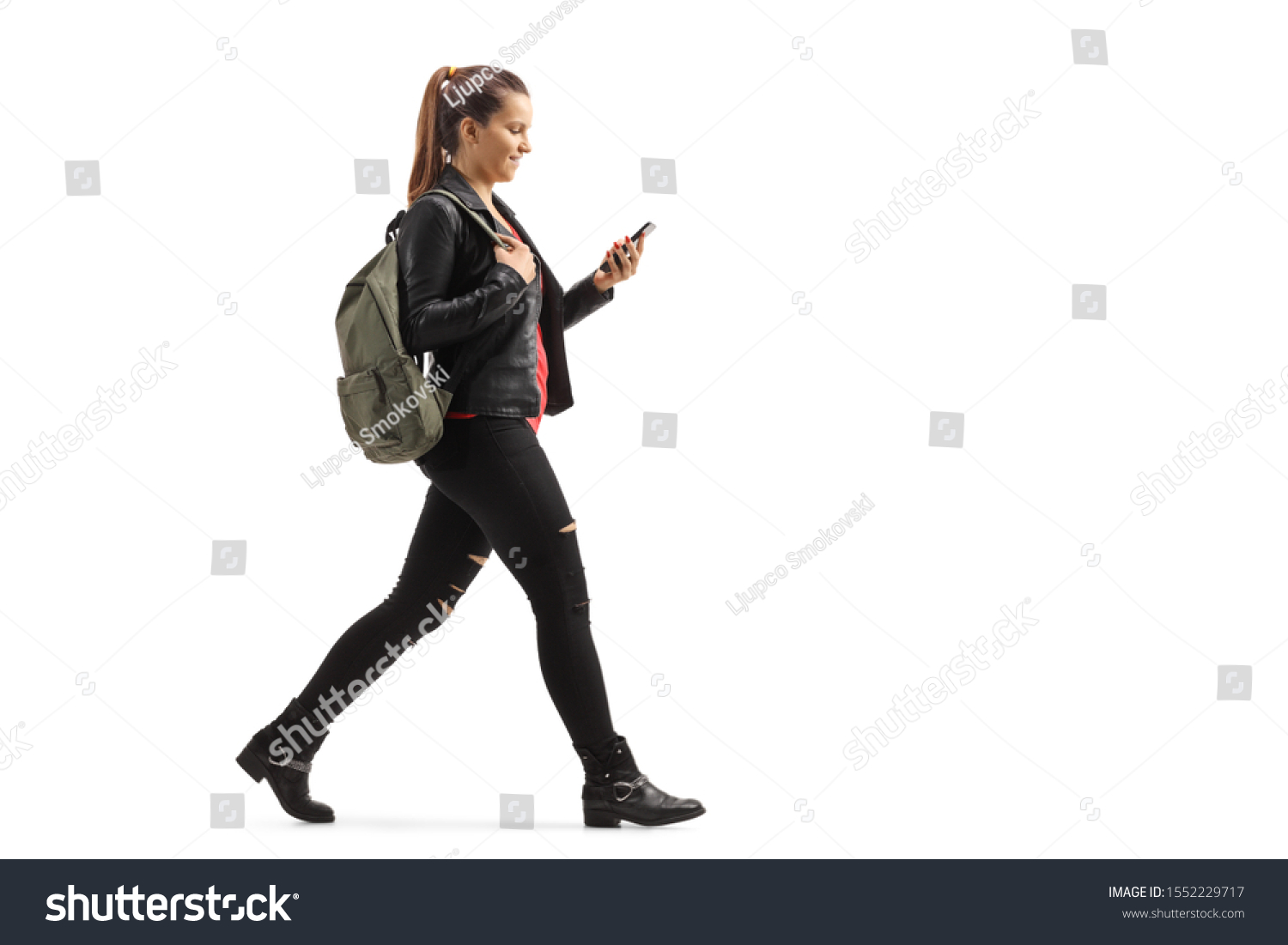 Full length profile shot of a girl with a backpack walking and looking at her mobile phone isolated on white background #1552229717