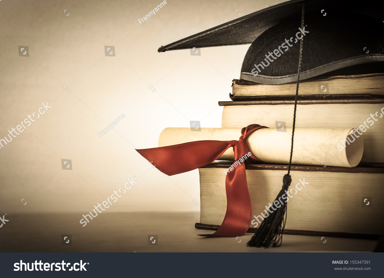 A mortarboard and graduation scroll, tied with red ribbon, on a stack of old battered book with empty space to the left.  Slightly undersaturated with vignette for vintage effect. #155347391