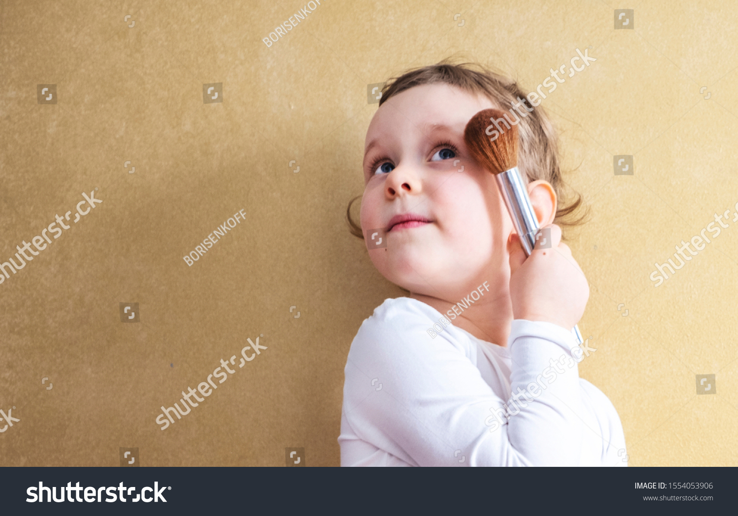 The child, a little girl of 4 years old, does makeup, imitates adults. A blonde with short hair in a white T-shirt holds a makeup brush in her hand. Orange wall and place to feast #1554053906