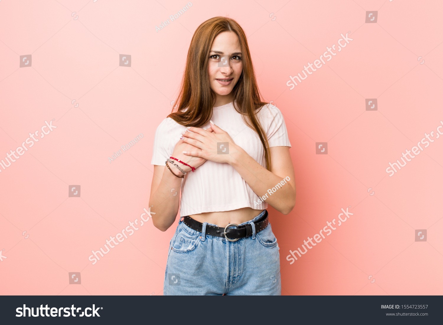 Young redhead ginger woman against a pink wall has friendly expression, pressing palm to chest. Love concept. #1554723557