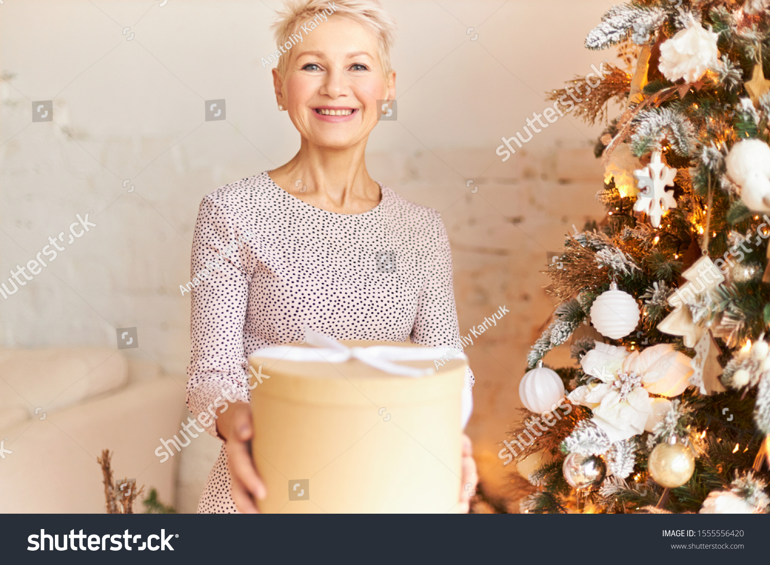 Gorgeous 50 year old European female wearing stylish dress wishing you Happy New Year, passing box with gift, looking at camera with radiant smile, being in festive mood. Holidays, festivity and party #1555556420