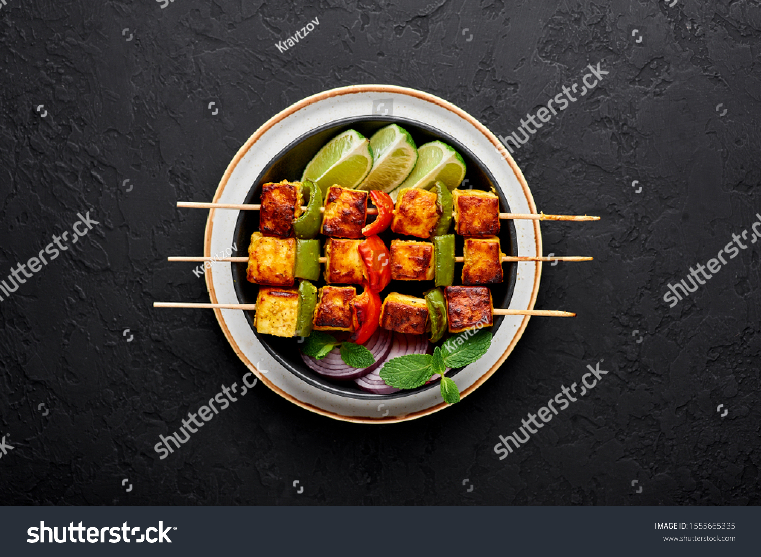 Paneer Tikka at skewers in black bowl at dark slate background. Paneer tikka is an indian cuisine dish with grilled paneer cheese with vegetables and spices. Indian food. Top view #1555665335