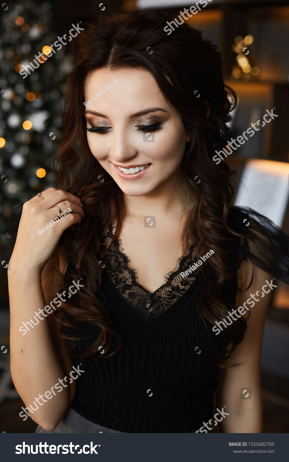 Portrait of a fashionable model girl with trendy hairstyle and trendy makeup in black blouse posing with festive lights of Christmas tree in background. Beautiful young woman with smooth perfect skin #1555682705