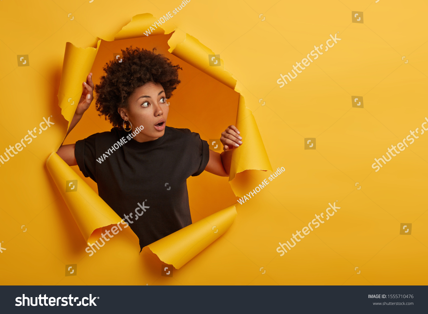 Photo of surprised Afro woman in black t shirt gasps from amazement, looks with scared face expression aside, dressed in black t shirt, unexpected to see something horrible, poses in torn paper wall #1555710476
