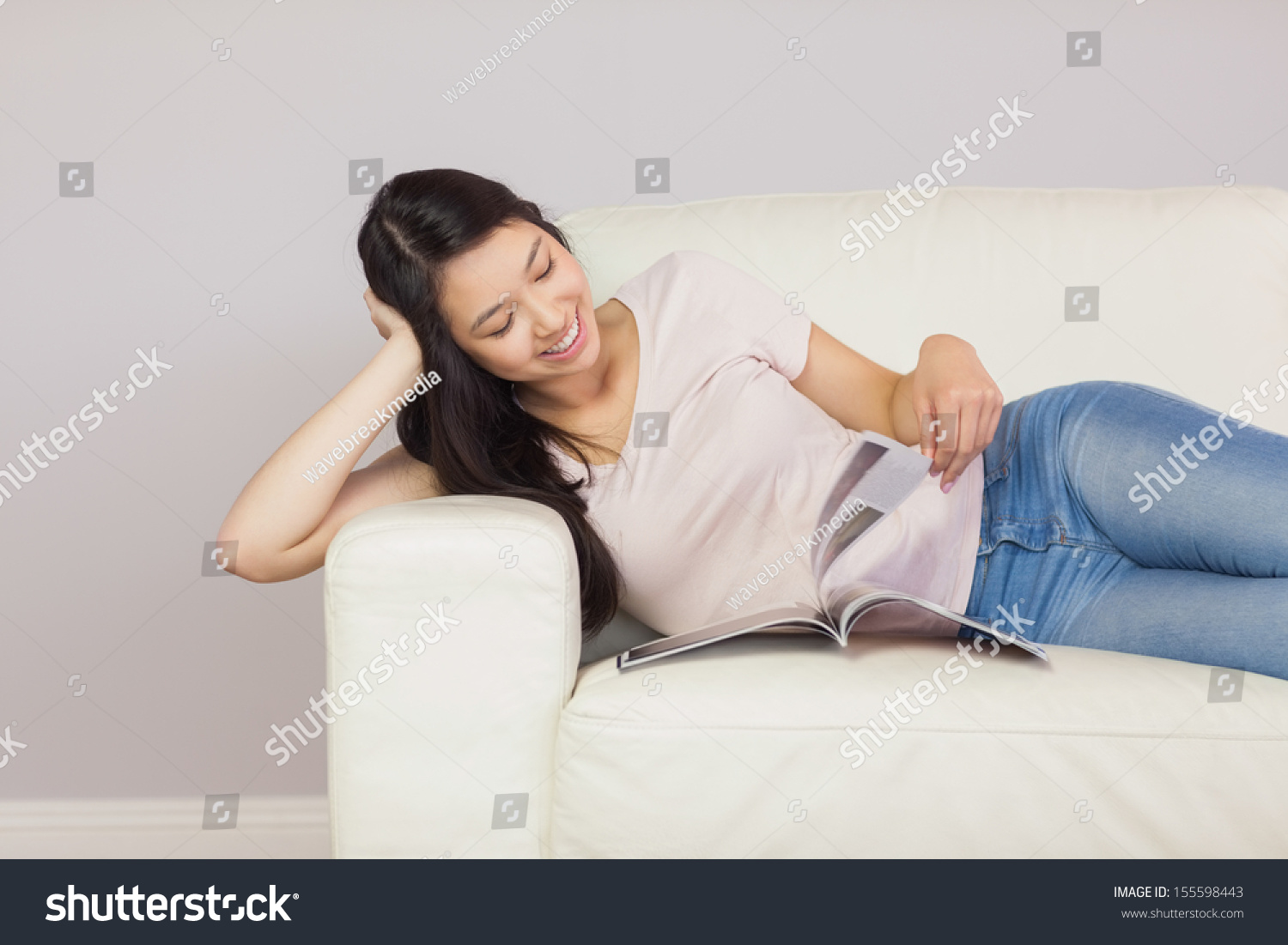 Cheerful asian girl lying on the sofa reading a magazine at home in the sitting room #155598443