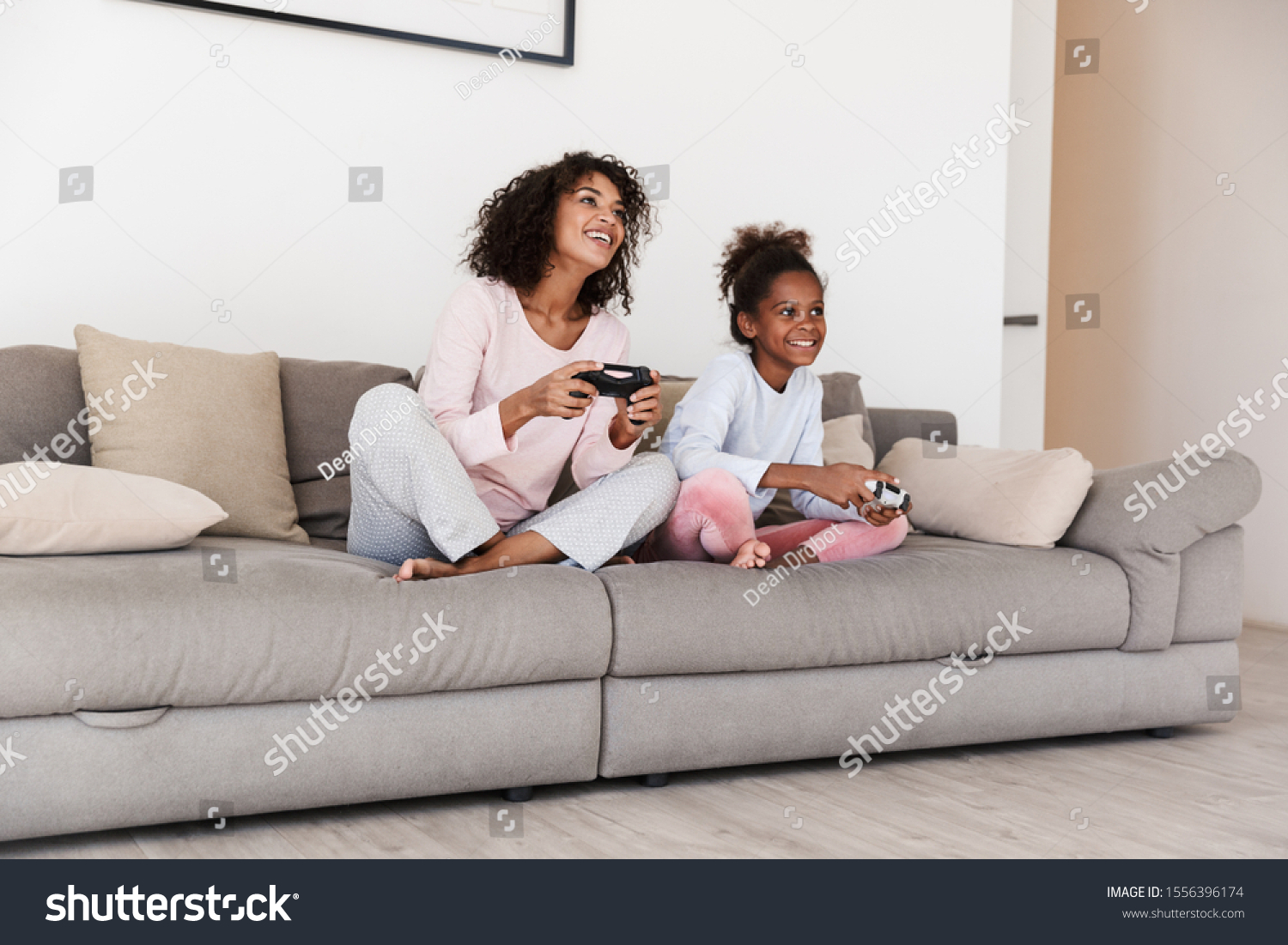 Smiling young mother and her little daughter wearing pajamas releaxing on a couch, playing video games #1556396174
