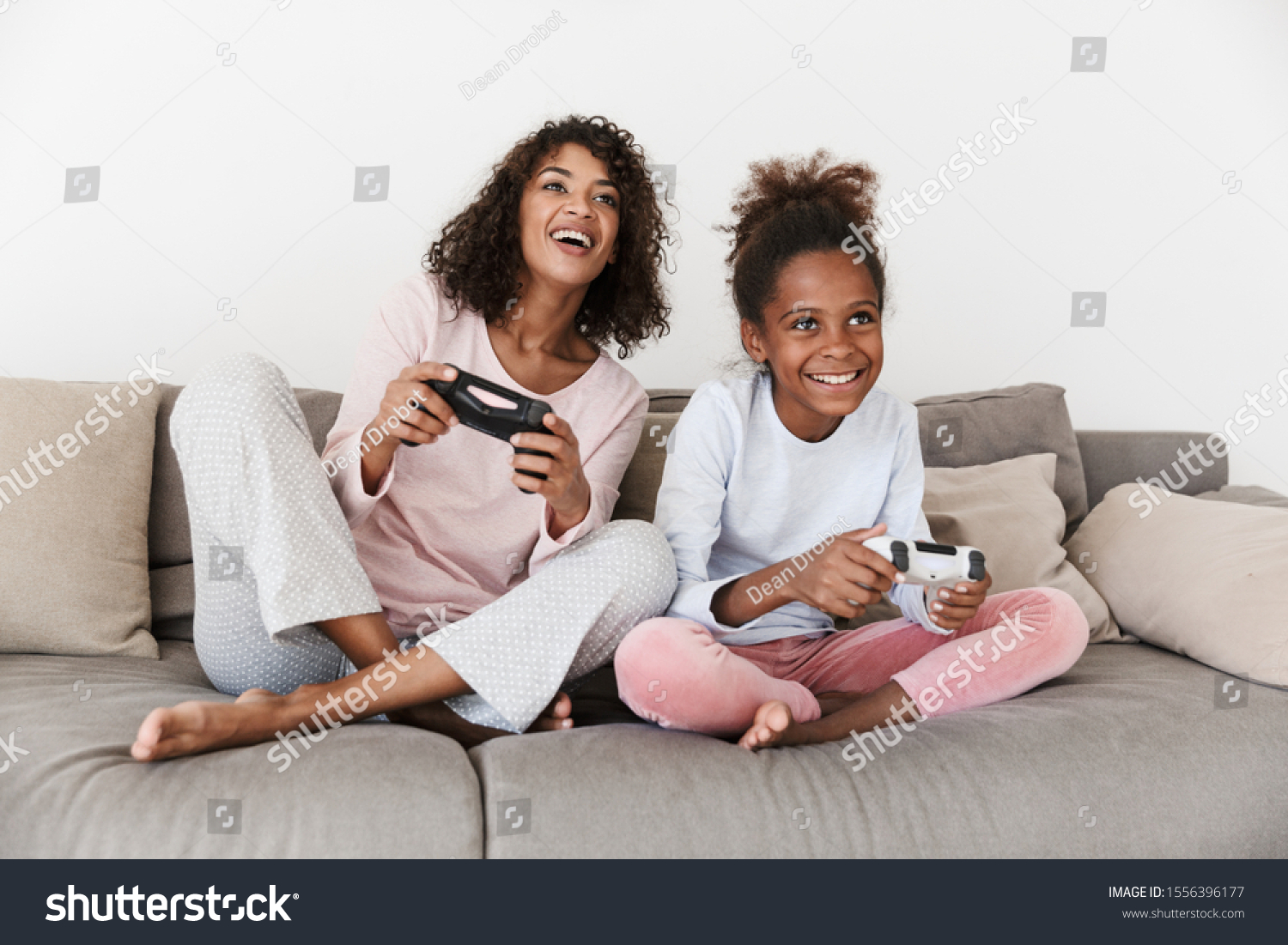 Smiling young mother and her little daughter wearing pajamas releaxing on a couch, playing video games #1556396177